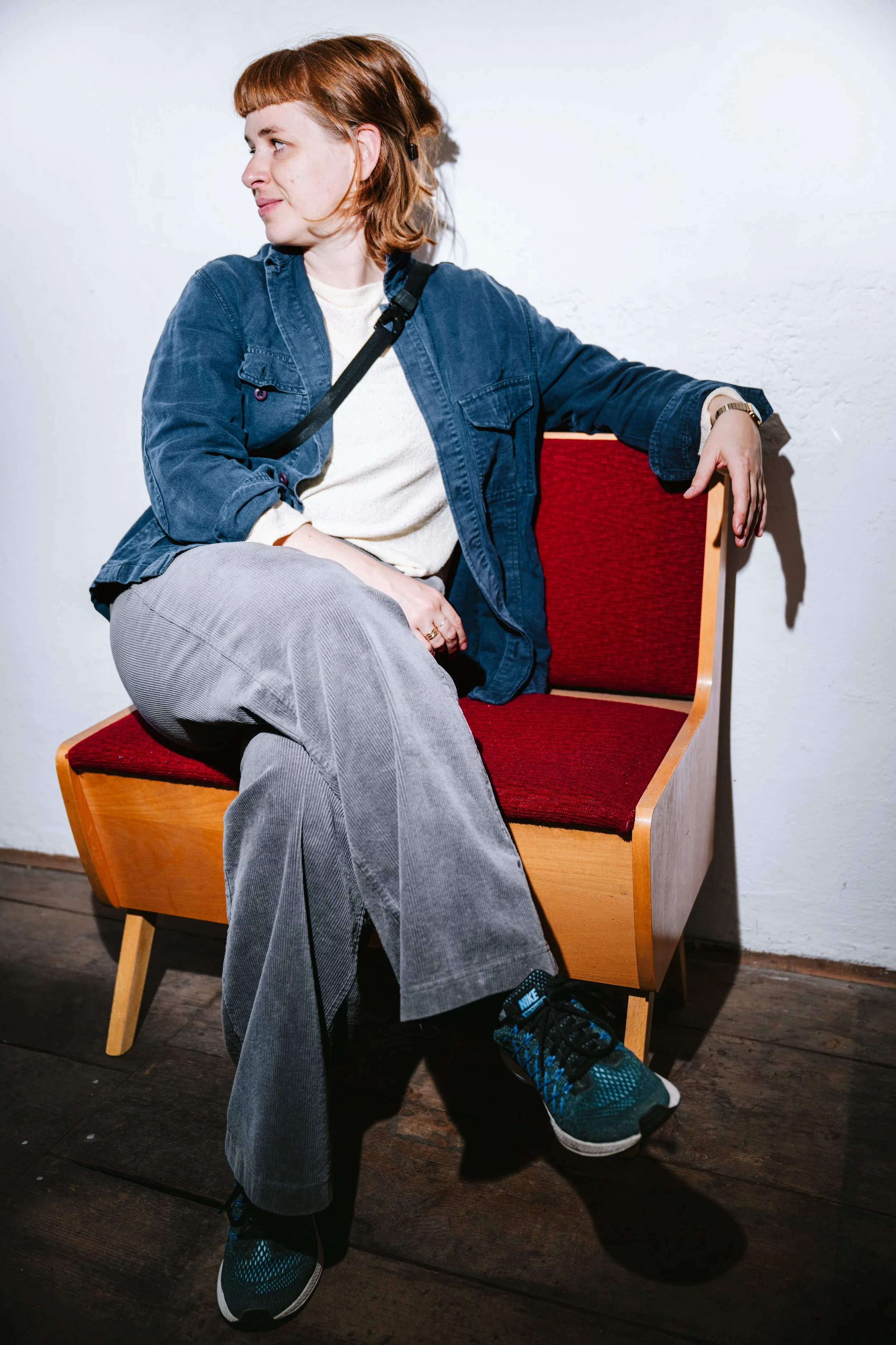 Junge Frau mit rotem Haar sitzt auf einem roten Sitz, blickt zur Seite, trägt eine blaue Jacke, graue Hose und blaue Sportschuhe, vor weißer Wand