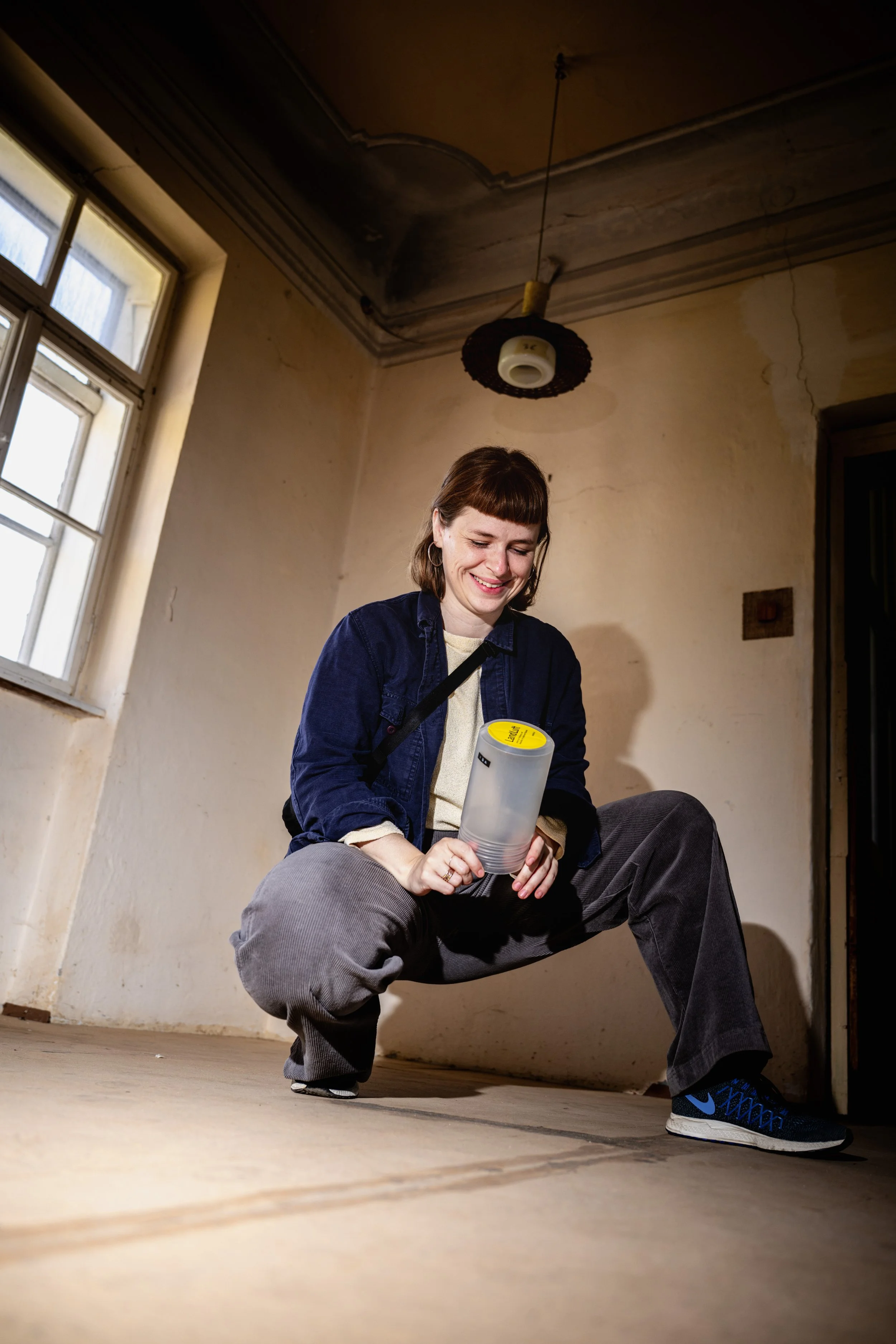 Eine junge Frau in einer ungewohnten Umgebung mit alten Wänden und einem breiten Fenster, die in die Hocke geht und eine transparente Box hält.