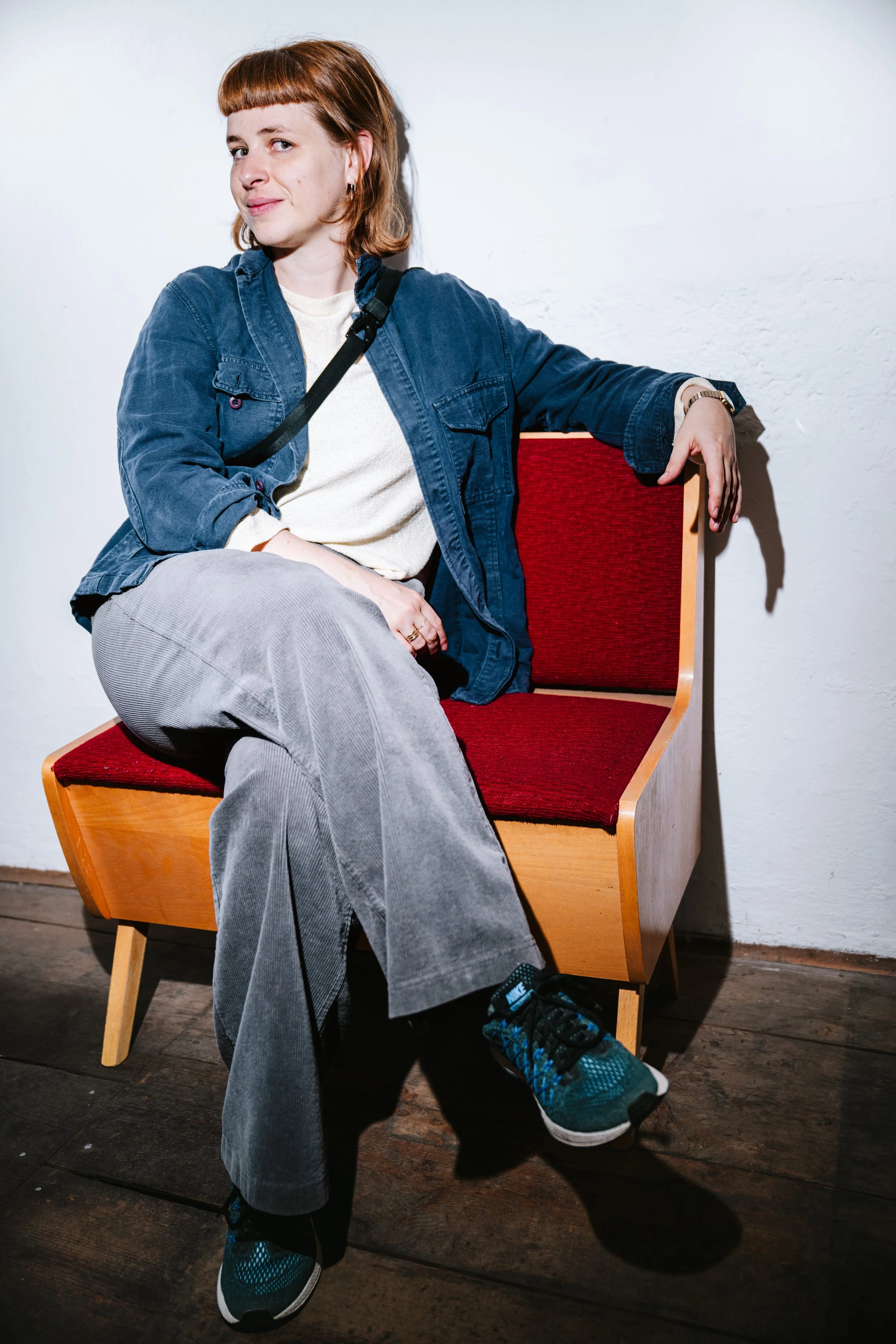 Junge Frau mit rotem Haar sitzt auf einem Holzstuhl mit rotem Sitzpolster, trägt eine dunkle Jacke, graue Hosen und sportschuhe, lehnt entspannt mit einem Arm auf der Rückenlehne, vor weißer Wand.