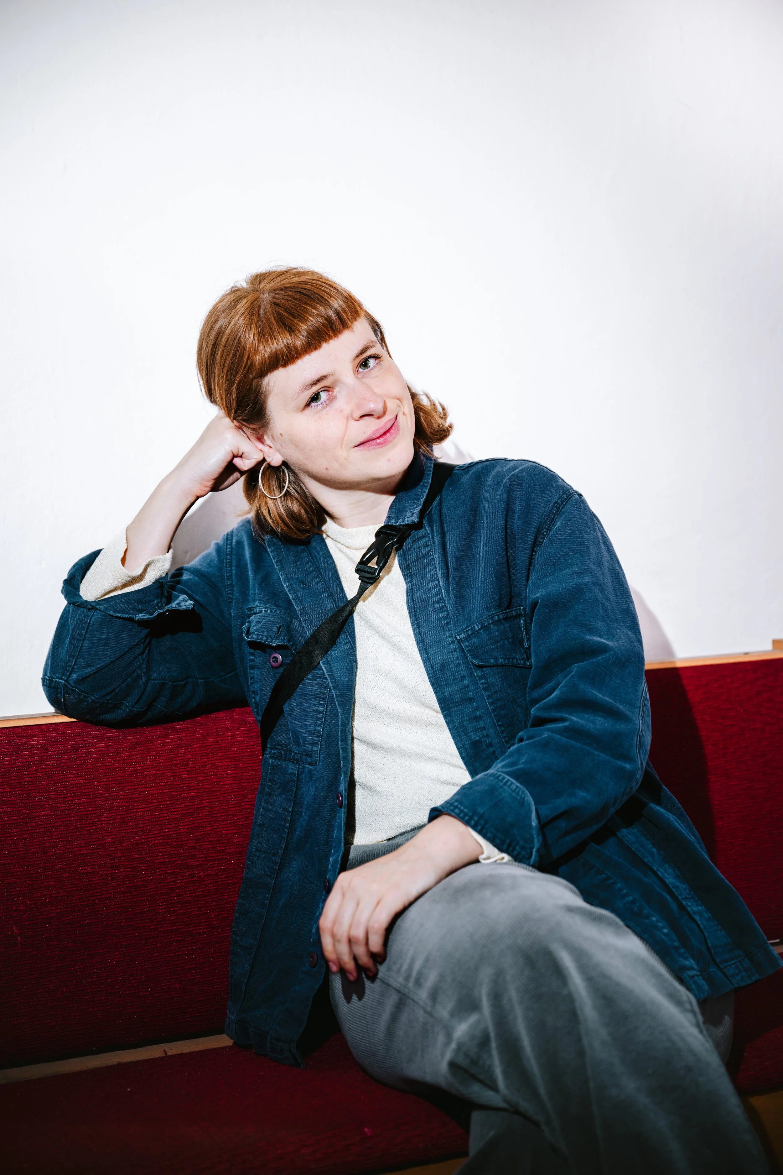 Eine junge Frau mit roten Haaren und Doppel-Ohrringen sitzt auf einer roten Couch, lehnt ihren Kopf an ihre Hand und blickt skeptisch.