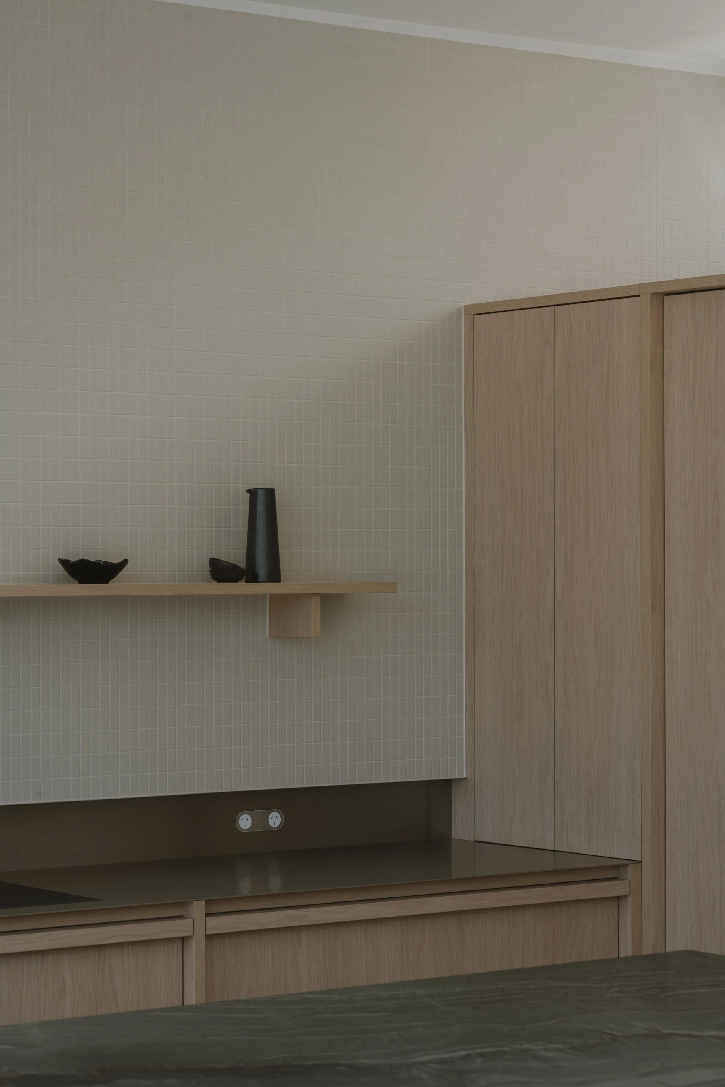 A minimalist kitchen corner with light wood cabinetry, a beige tiled wall, and a black vase with a small bowl on a floating shelf.