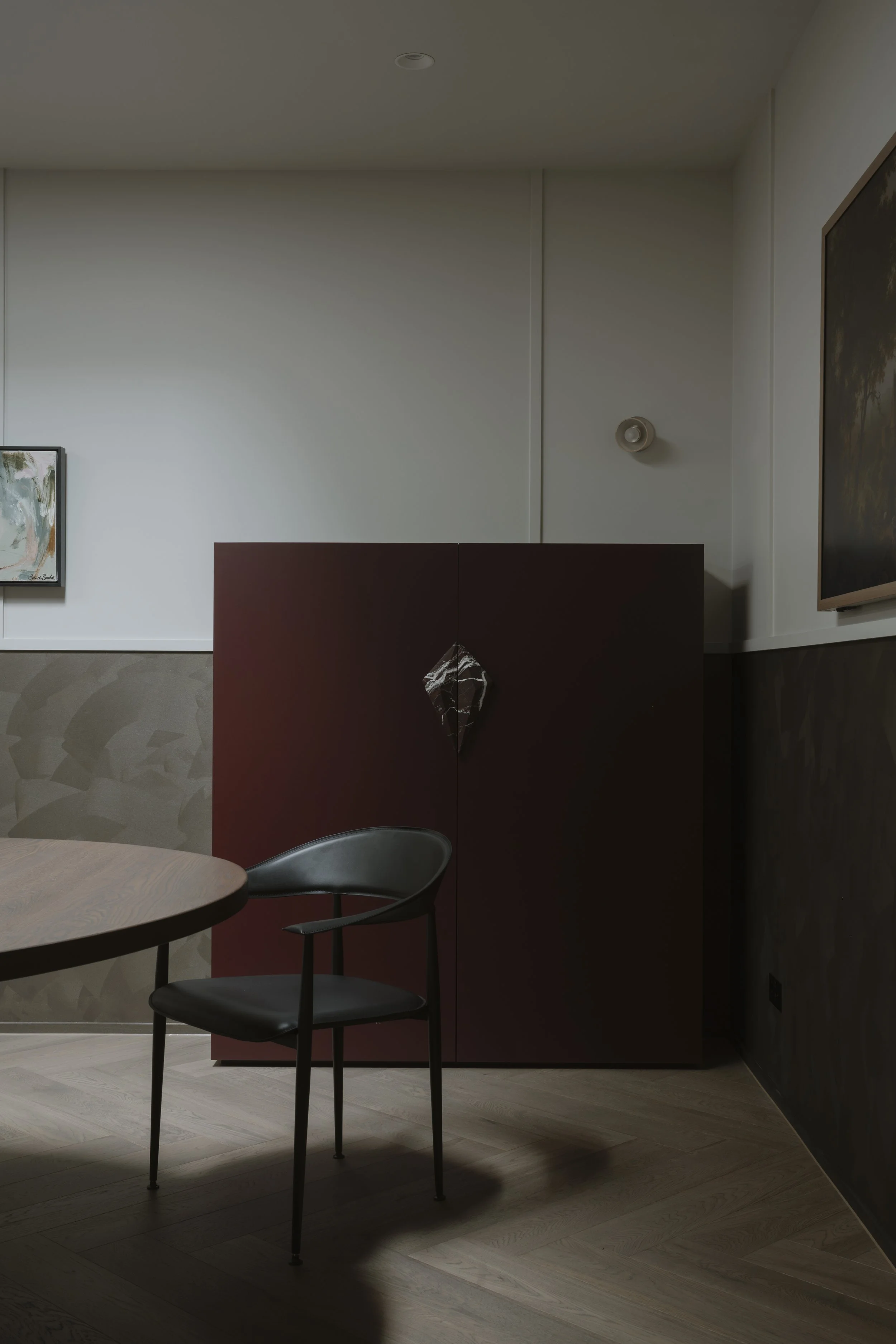 Interior of a modern room with a burgundy cabinet, a round wooden table, and a black chair. The room has white walls, artwork, and subtle lighting.