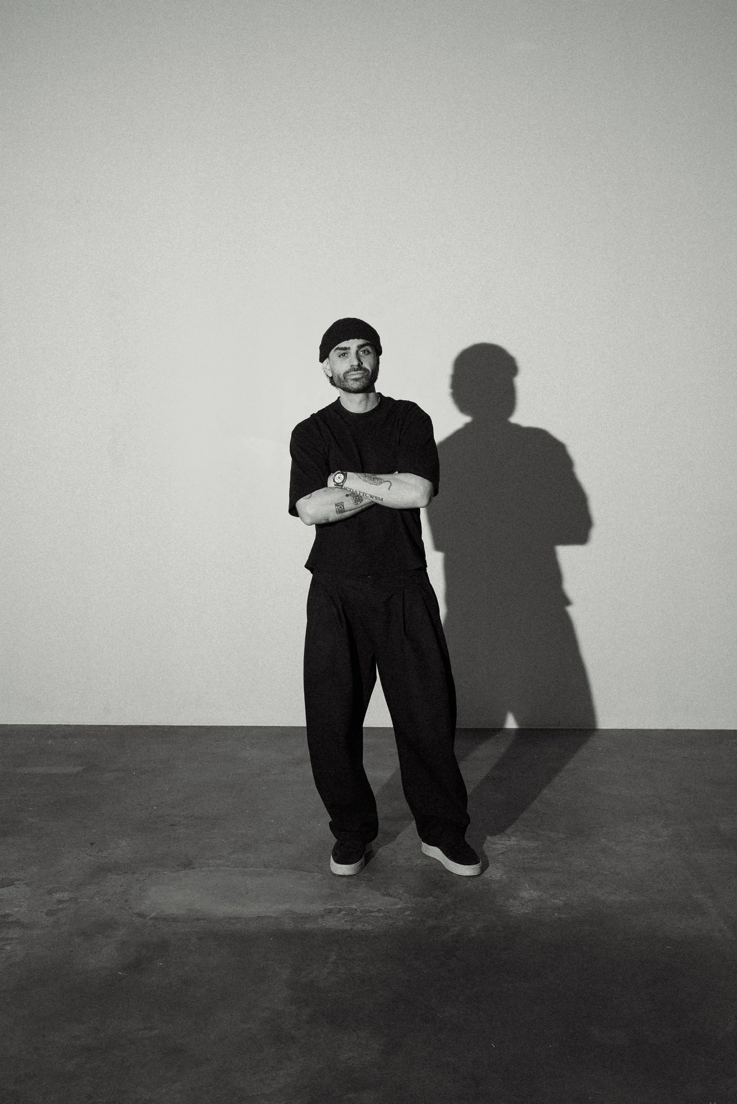 Black and white photo of a man sitting on a chair against a plain wall, with one arm resting on his knee and the other hanging down. The shadows cast on the wall behind him show his profile.