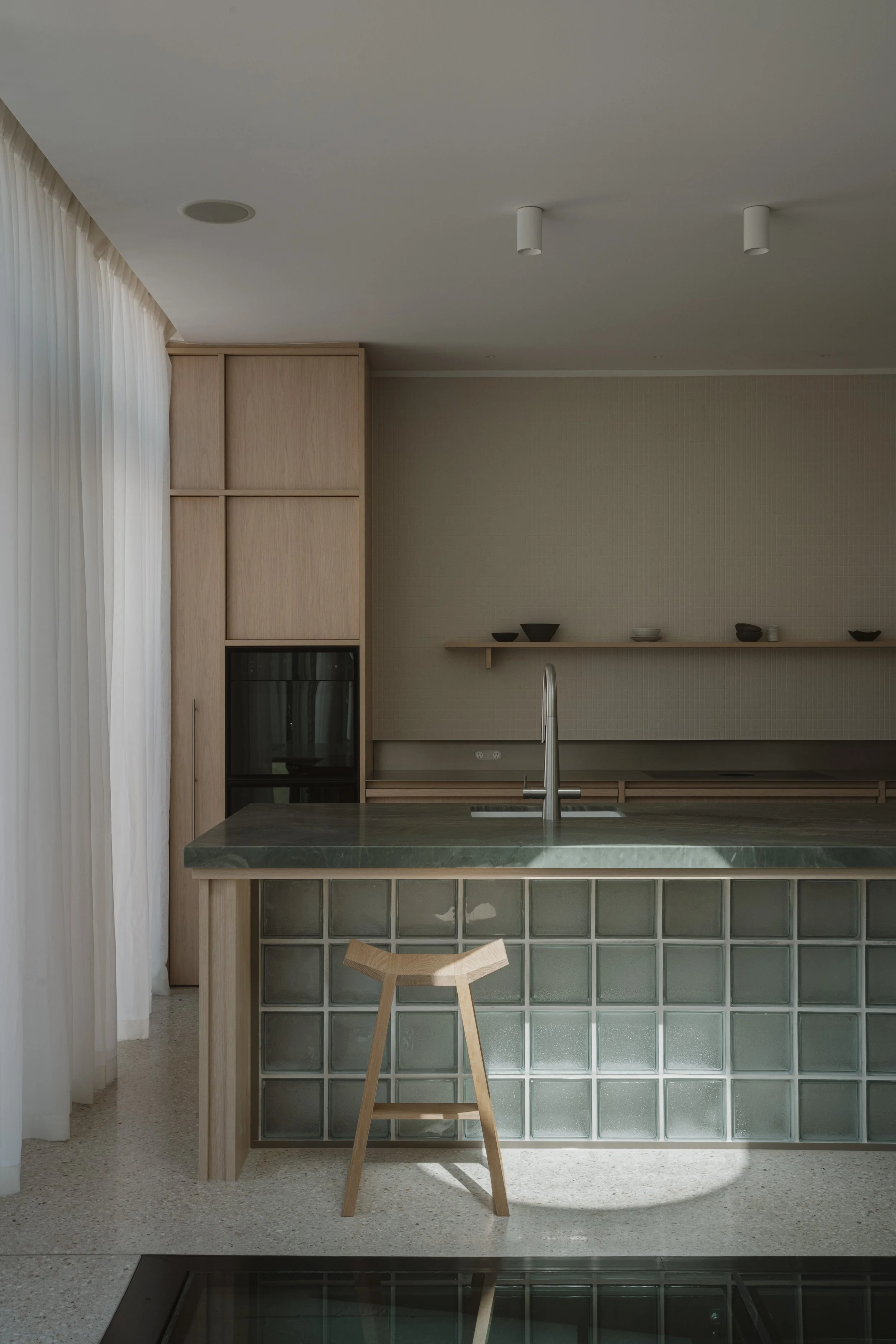A modern kitchen with a marble island, a wooden stool, and a glass brick wall. There is a shelf with bowls and a curtain on the left side.