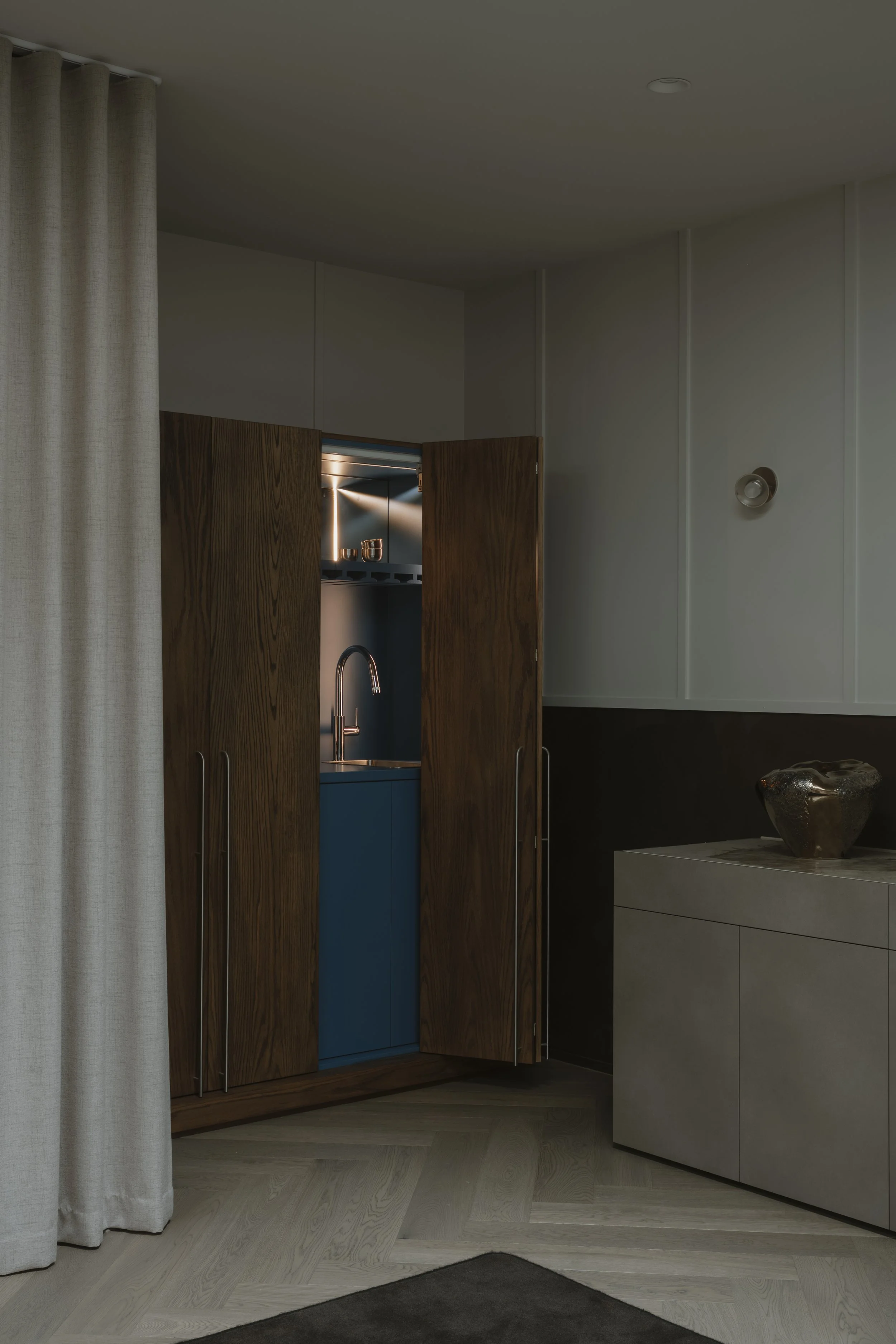 Interior view of a modern kitchen with wooden and blue cabinets, a stainless steel sink with a curved faucet, and a glimpse of a stove in the background. Light-colored wood flooring and a beige curtain on the left side. A beige cabinet with a metalli