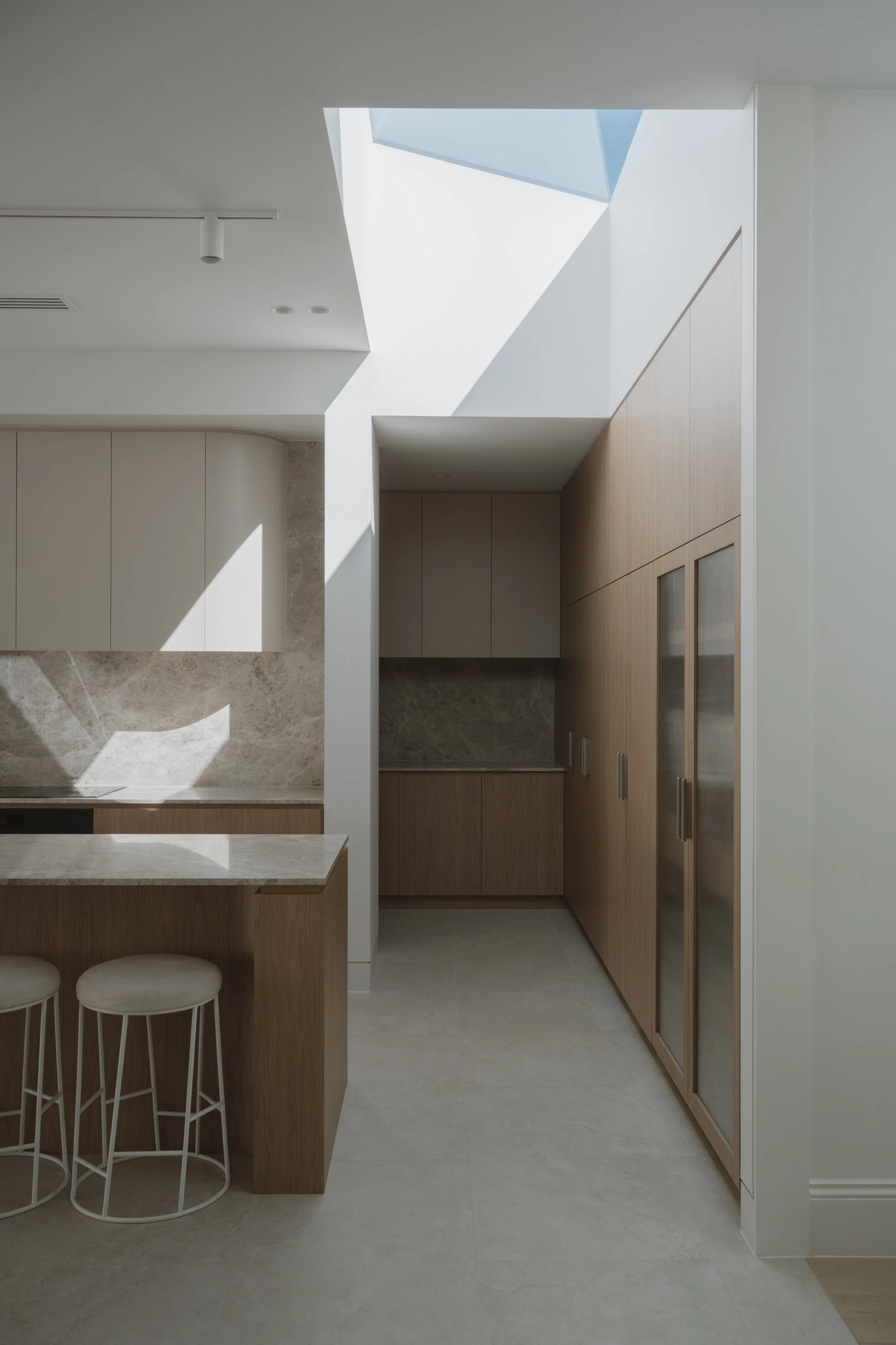 Modern kitchen with minimalist design featuring wooden cabinets, a marble countertop, an island with bar stools, and a skylight allowing natural light to illuminate the space.