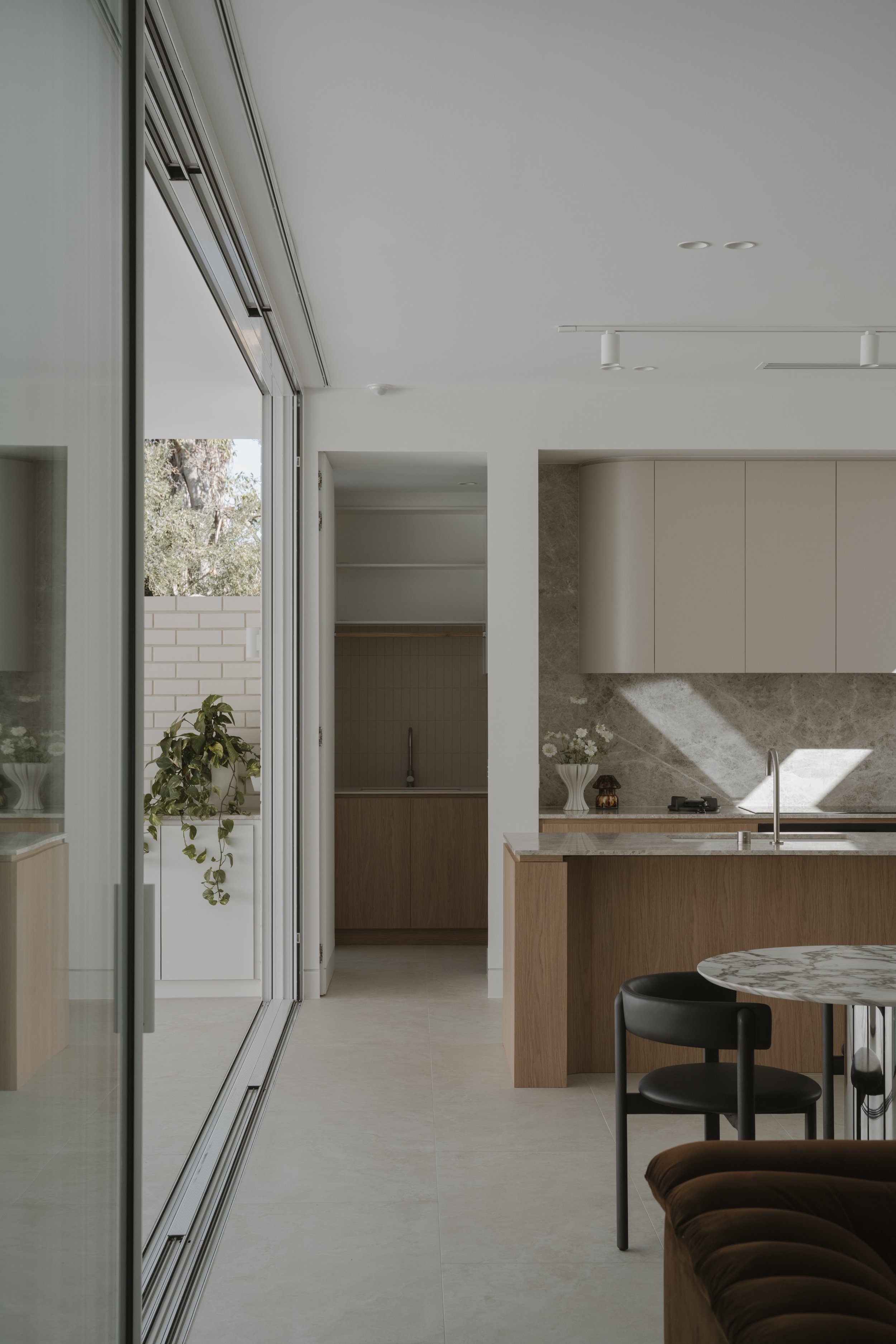 Modern kitchen with light-colored cabinets, marble countertop island, and seating area. Large windows allow natural light, with outdoor trees and brick wall visible.
