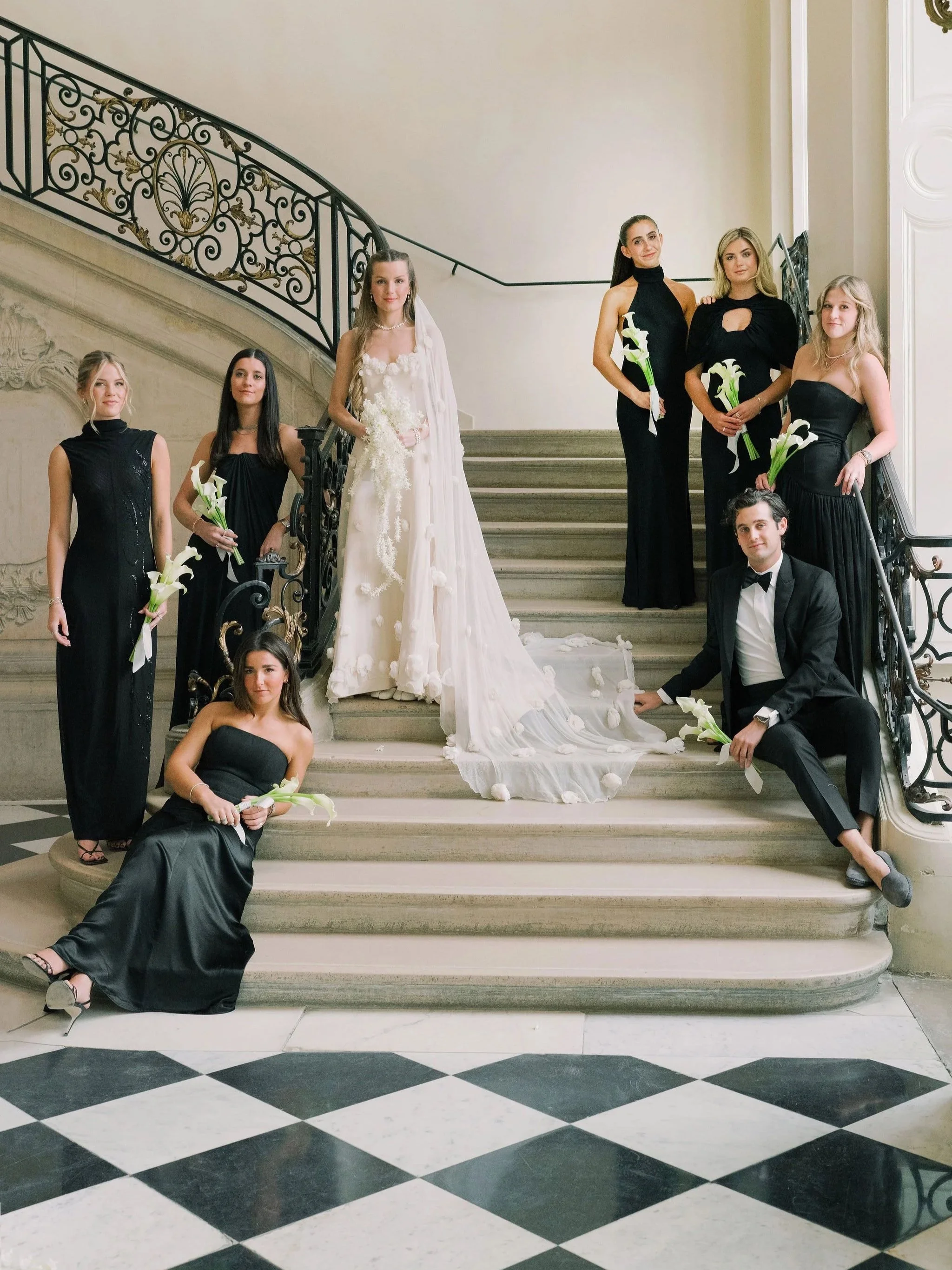 Bride and bridesmaids in black gowns on the grand staircase of the Musée Rodin in Paris, captured as part of a wedding produced by The Petaler Floral & Events Co., luxury wedding planner for American couples getting married in France.