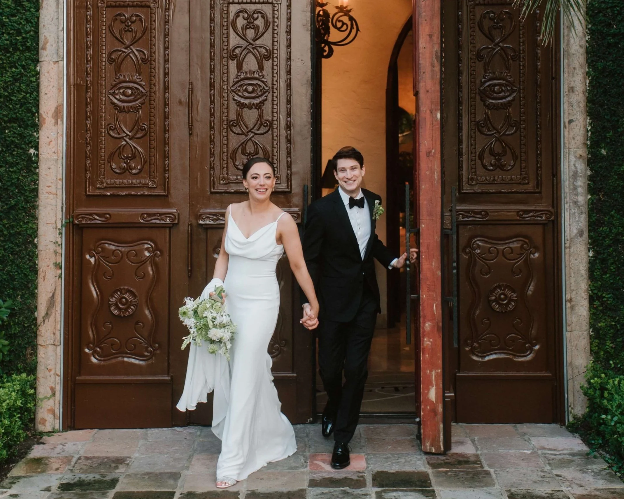 Bride and groom holding hands as they step through ornate wooden doors following their wedding ceremony, the bride carrying a green and white bouquet in a refined architectural setting.