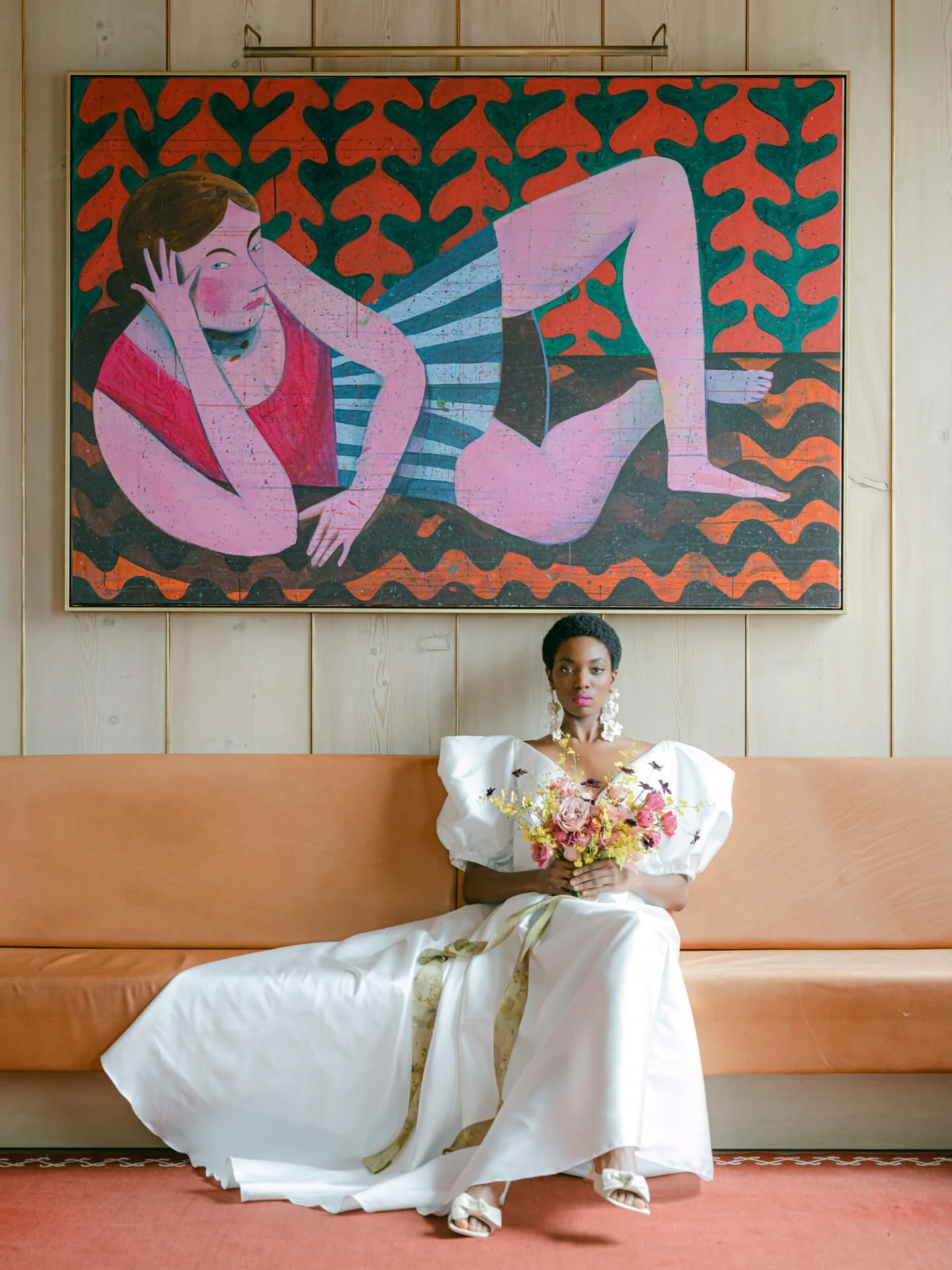 Bride in a white gown holding a colorful garden-style bouquet of pink, yellow and burgundy flowers beneath a vibrant modern painting.