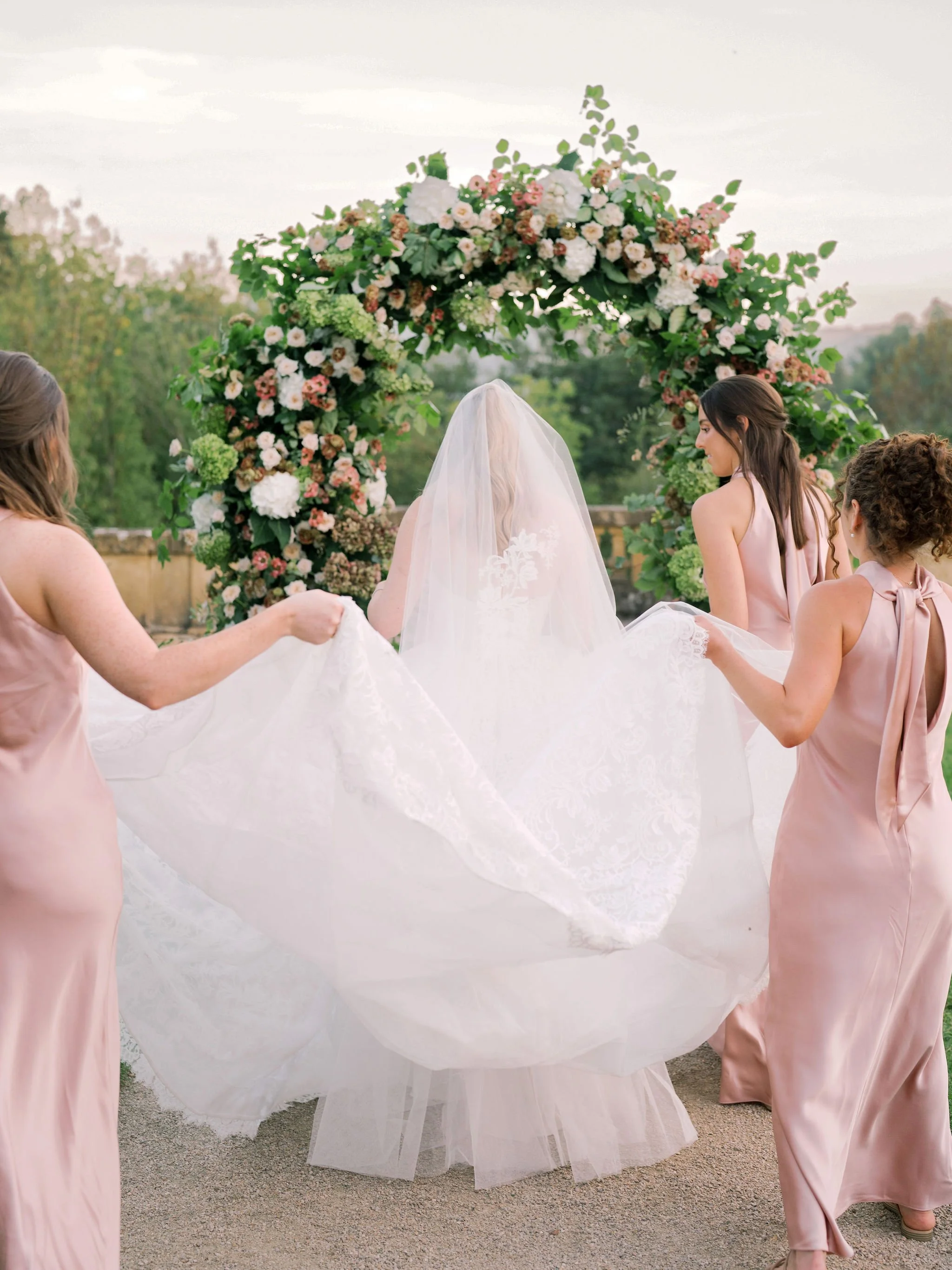 Bride walking toward a lush garden-style floral arch at Domaine des Halles, with her bridesmaids lifting her lace train, styled by The Petaler Floral and Events Co., wedding planner in France