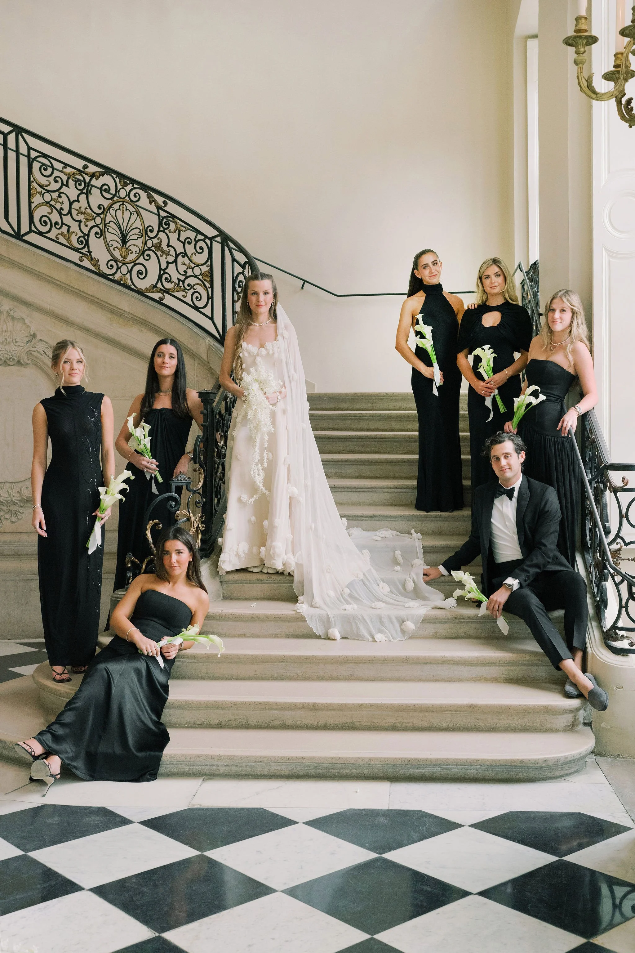 Bride in an elegant white wedding gown surrounded by her bridal party on the grand staircase at Musée Rodin in Paris, France. Bridesmaids wear black gowns and carry minimalist white calla lily bouquets.