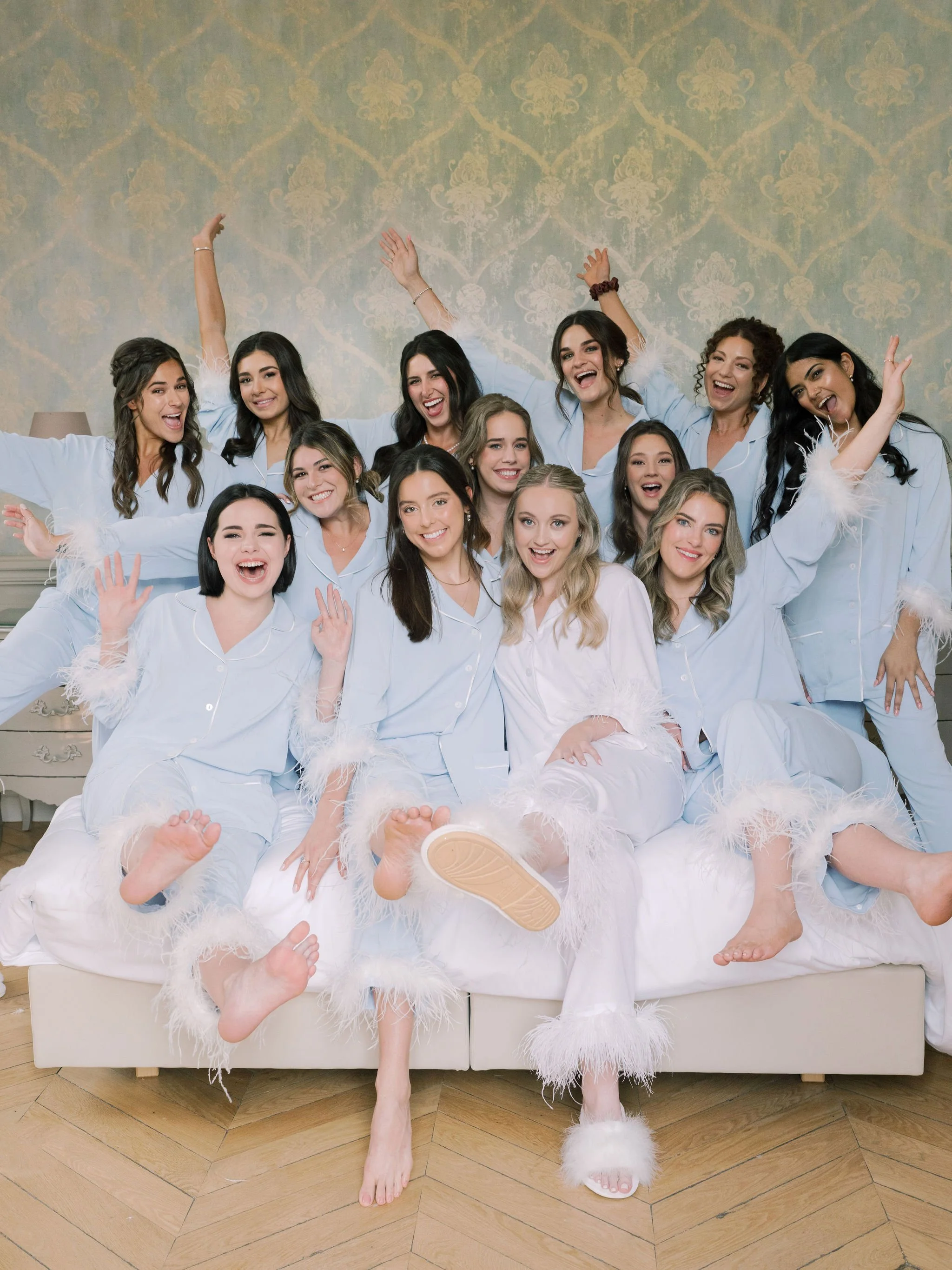 Bride and bridesmaids in feather-trimmed pajamas celebrating together on the wedding morning at Domaine des Halles, styled by The Petaler Floral and Events Co., wedding planner in France.