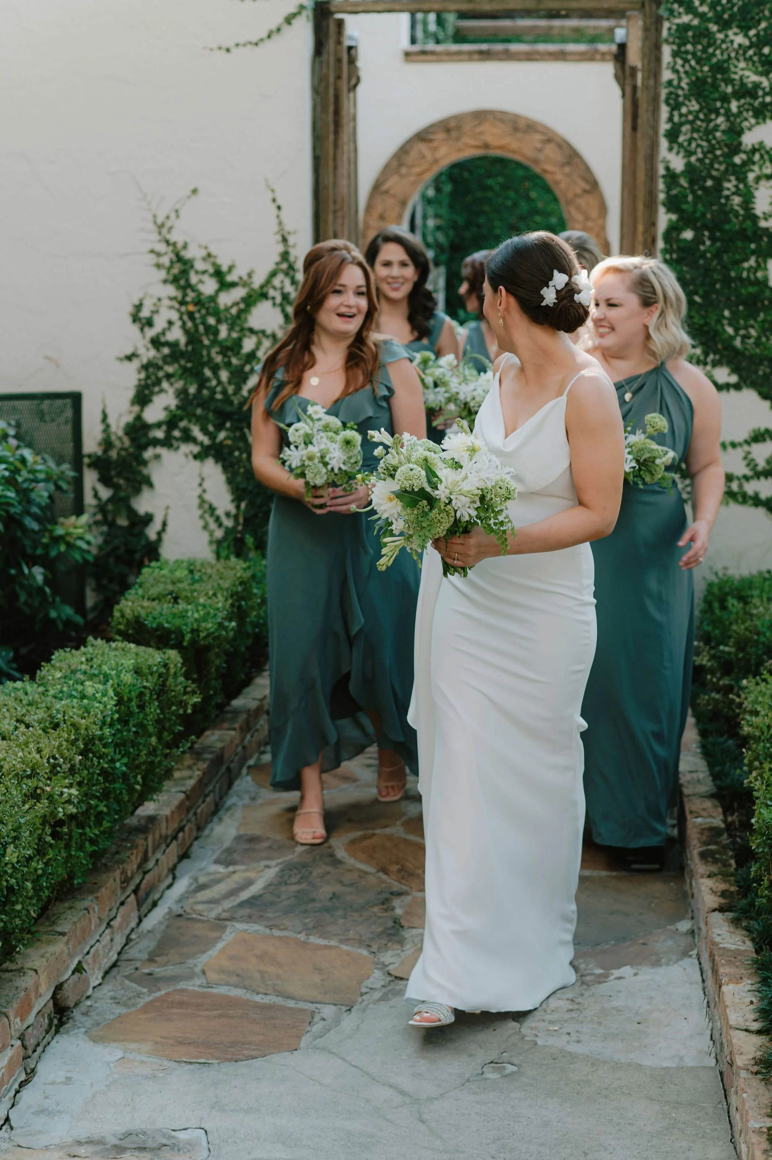 Bride and bridesmaids walking through a garden path, styled in soft green tones and carrying refined green and white floral bouquets in a contemporary wedding setting.