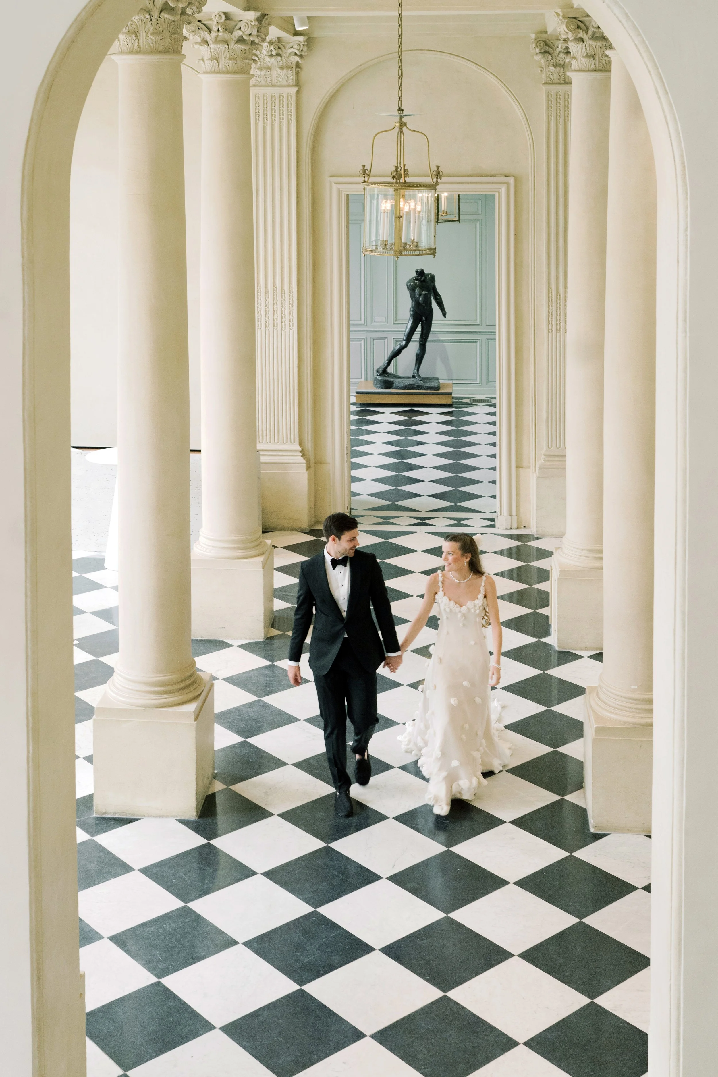Bride and groom walking through a grand black-and-white gallery at the Musée Rodin. Wedding planing and design by The Petaler Floral & Events Co., specialists in wedding productions for American couples marrying in France.