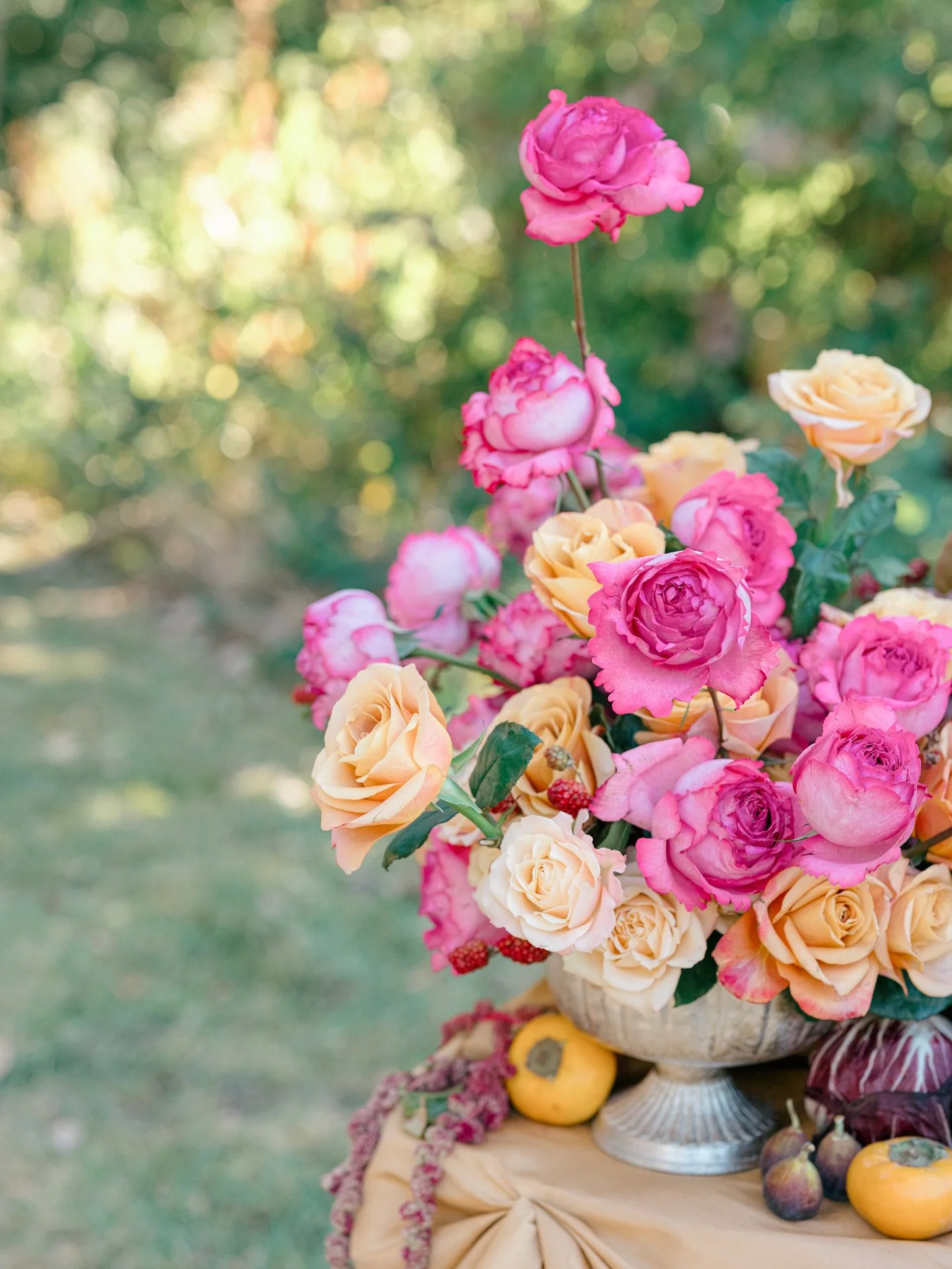 Peach and pink garden roses arranged in a stone urn with figs and citrus, styled as an outdoor still life.