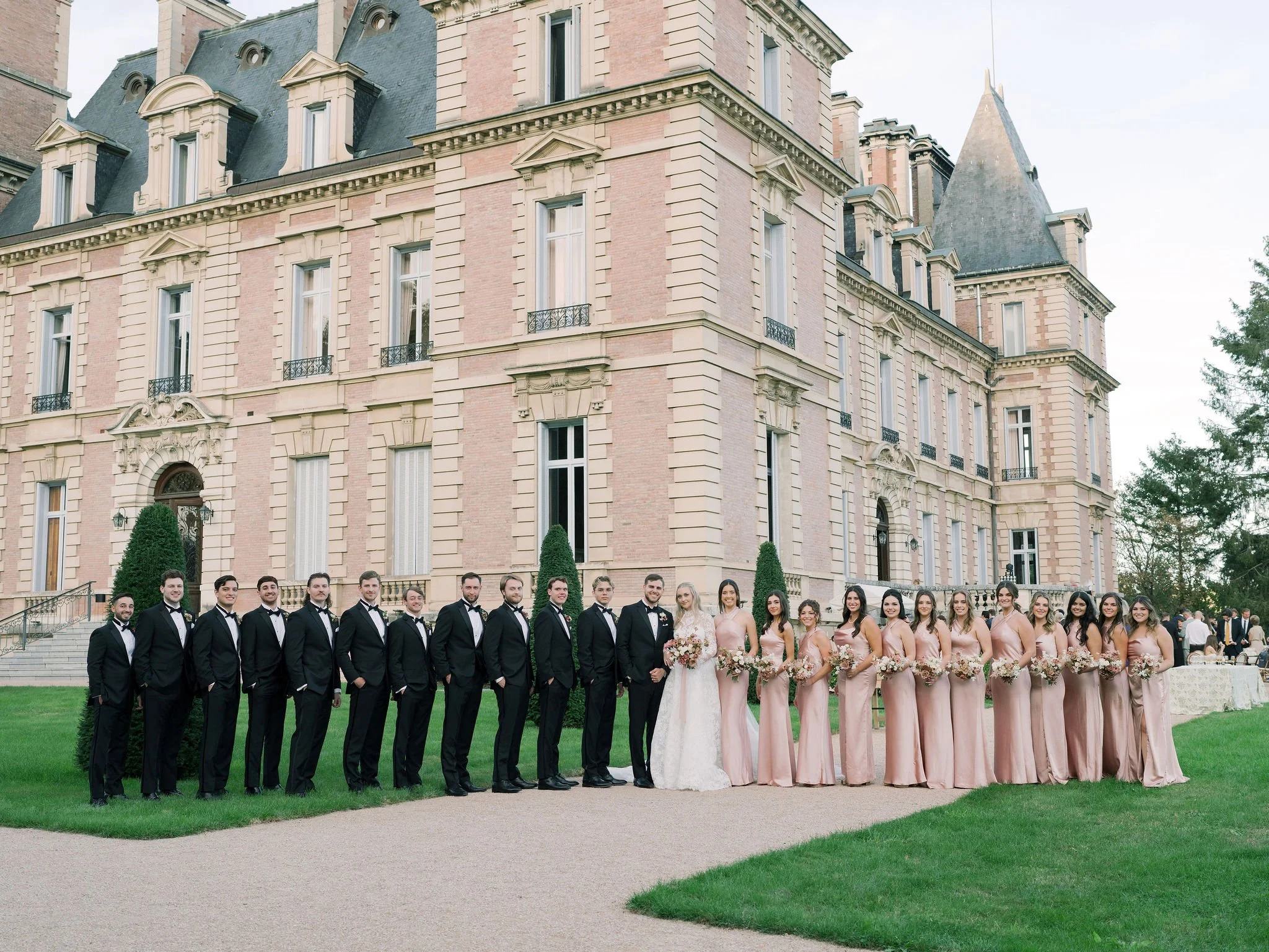 Wedding party portrait in black tie and blush gowns in front of the grand façade of Domaine des Halles, with florals by The Petaler Floral and Events Co., wedding planner in France.