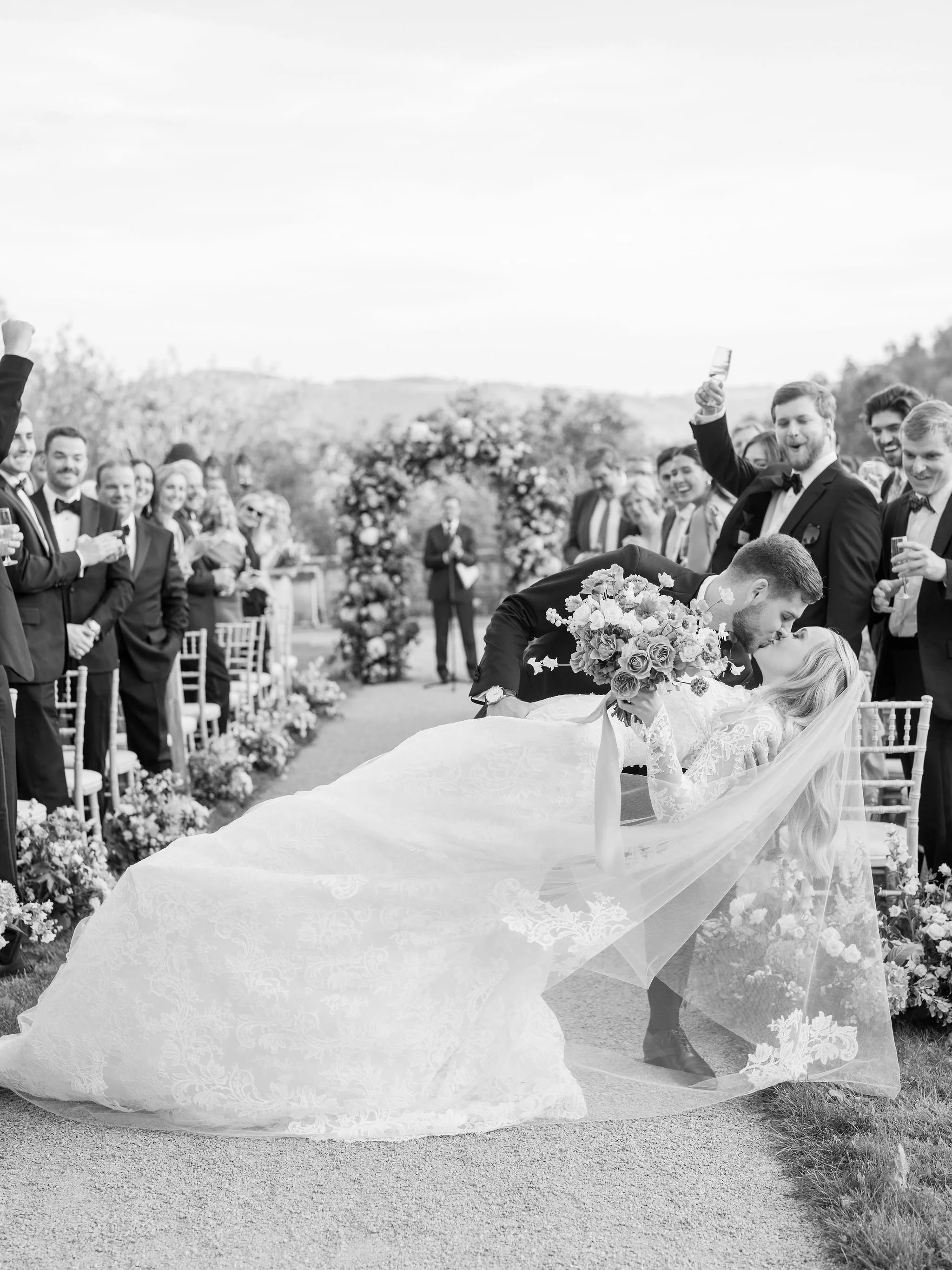 Couple sharing a celebratory kiss during their ceremony recessional beneath a garden-style floral arch at Domaine des Halles, surrounded by cheering guests, with florals by The Petaler Floral and Events Co., wedding planner in France.