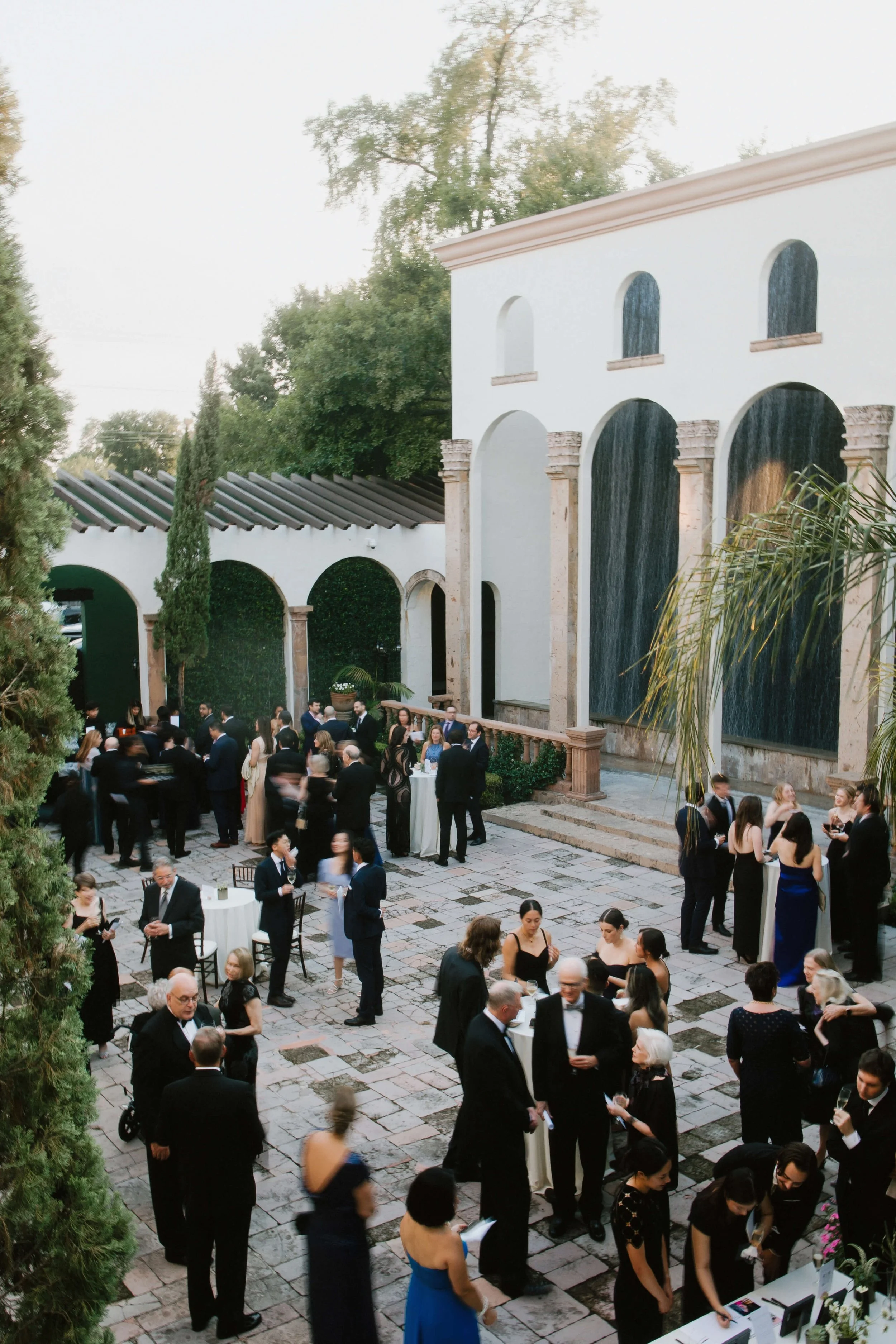 Wide view of a lively wedding cocktail hour unfolding in a historic courtyard, with guests mingling beneath arches and palm trees in a refined architectural setting.