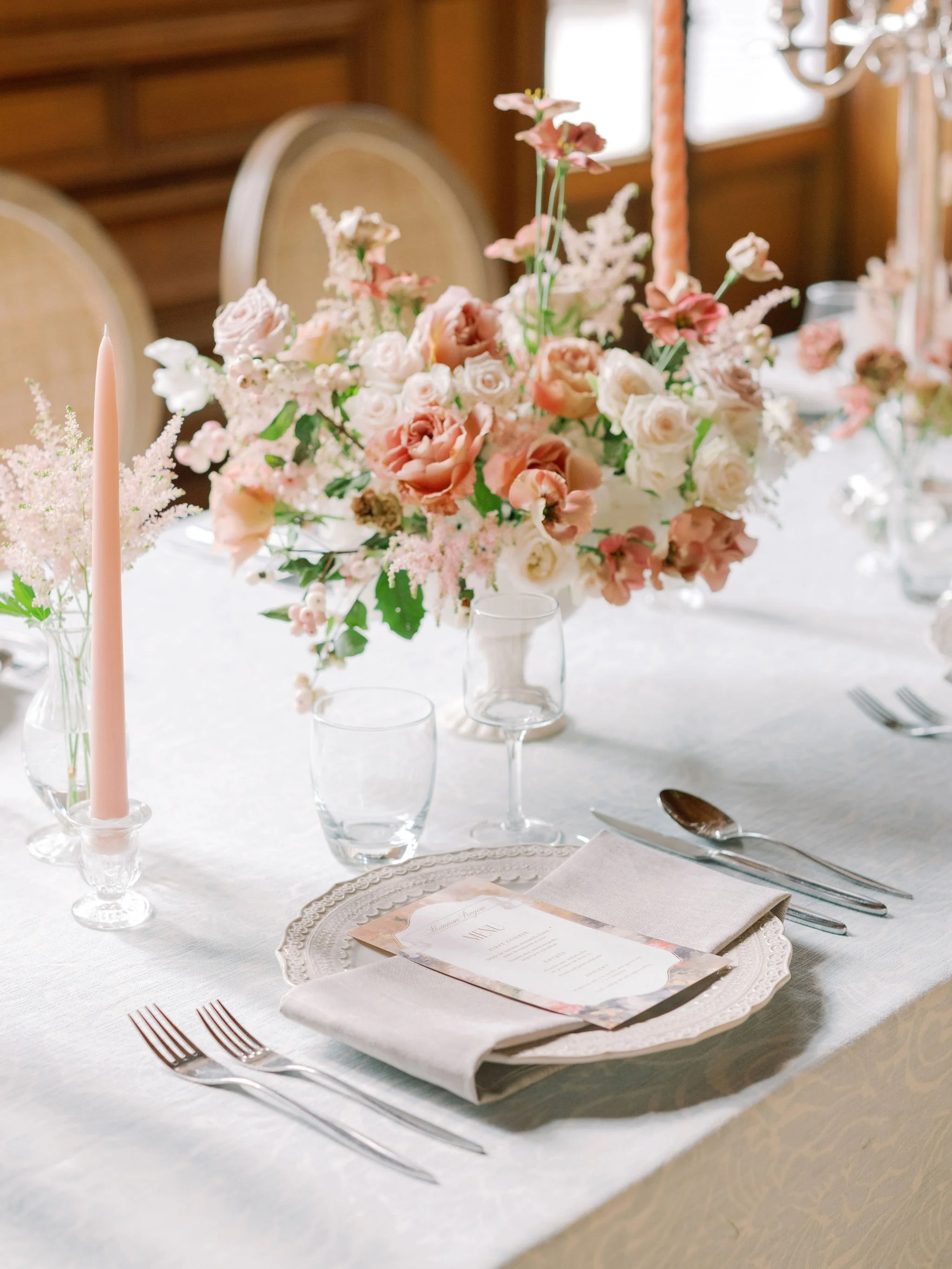 Elegant place setting with linen napkin, menu card, blush taper candles, and garden-style floral centerpiece at a wedding reception at Domaine des Halles, designed by The Petaler Floral and Events Co., wedding planner in France.