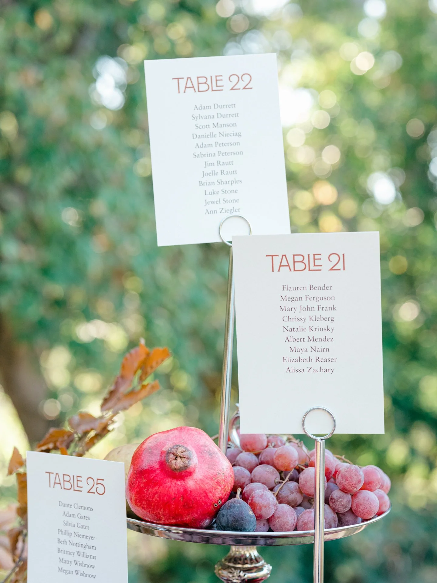 Wedding escort card display with modern table cards and seasonal fruit styled on a silver compote in an outdoor garden setting.