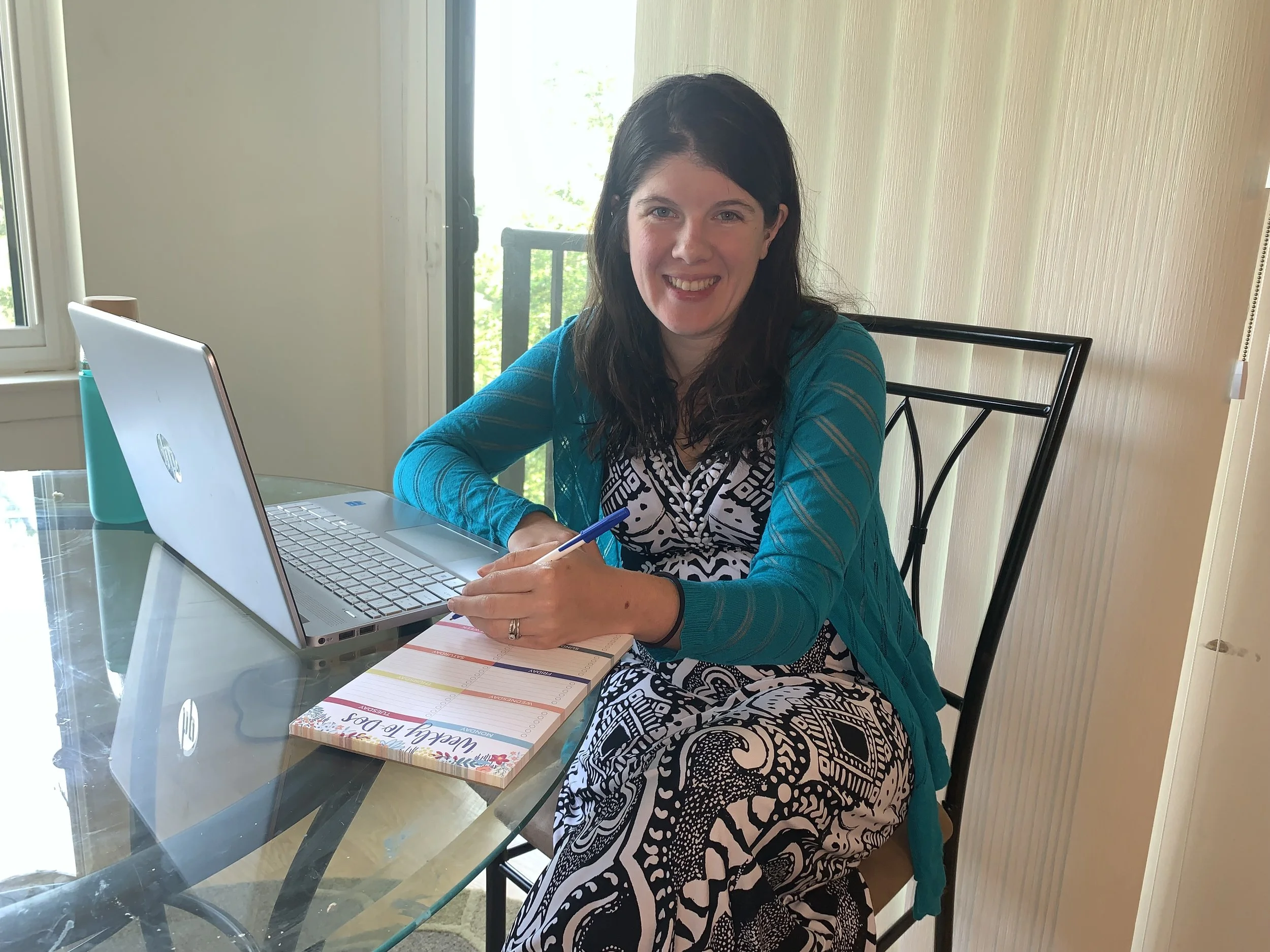 Founder of Stunning Jade, brunette with long hair. She is wearing a black and white dress with a blue shawl. With computer, notepad and pen.