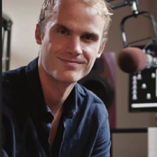 A man with short blonde hair smiling in a recording studio, sitting in front of a microphone and audio equipment.