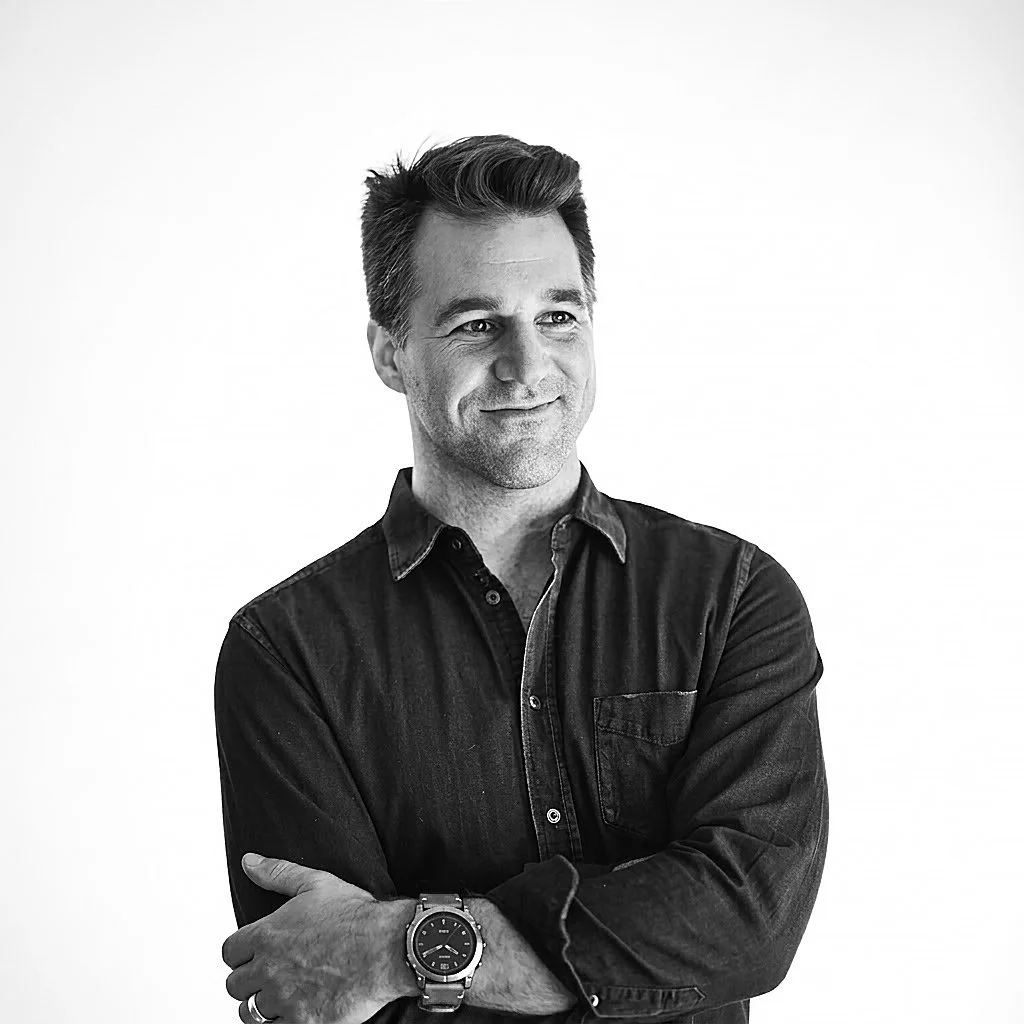 A black-and-white photo of a man with styled hair, wearing a dark button-up shirt with rolled sleeves, a wristwatch, and a ring, standing against a plain background, with arms crossed and a slight smile. This is photographer Michael Khachadoorian