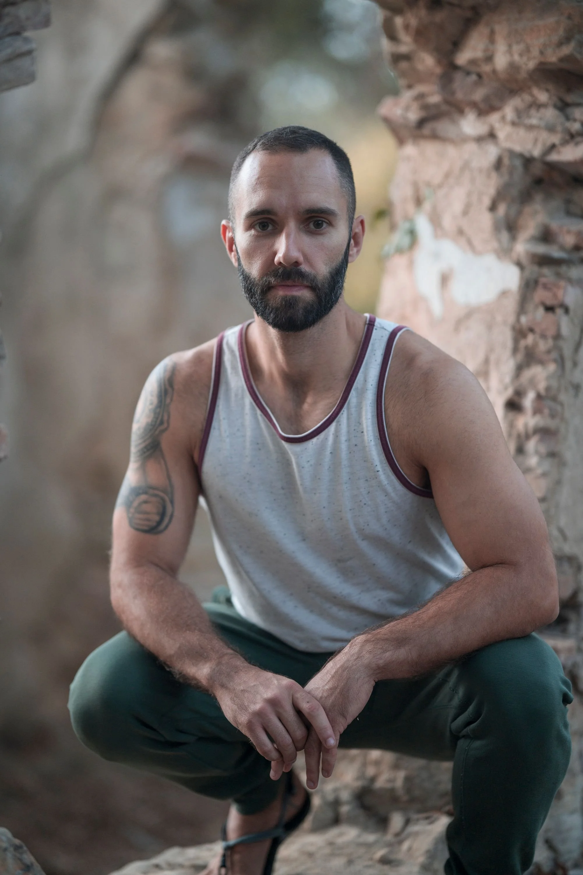 A man with a beard, short dark hair, wearing a white sleeveless shirt with maroon trim and dark green pants, is squatting outdoors between stone ruins with a blurred natural background.