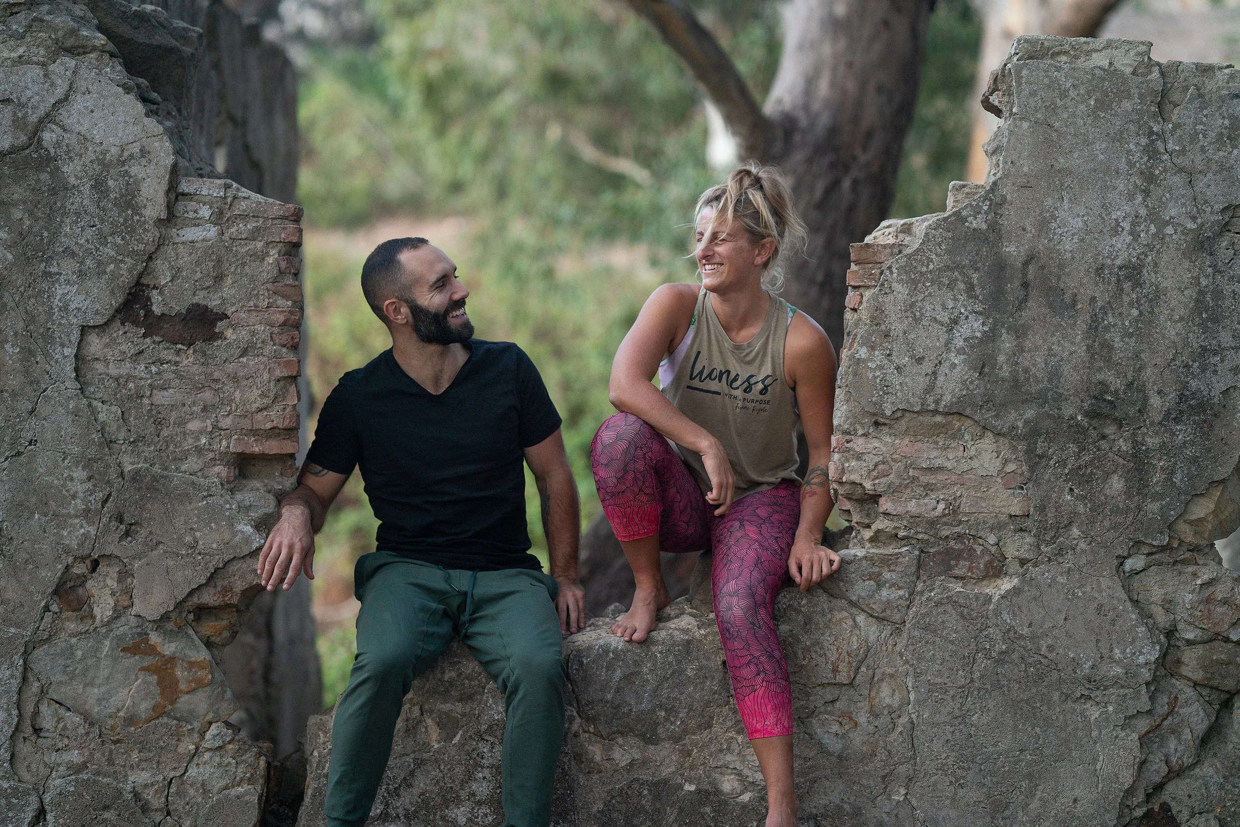 A man and woman sitting on a large rock, smiling and looking at each other, surrounded by trees and nature.