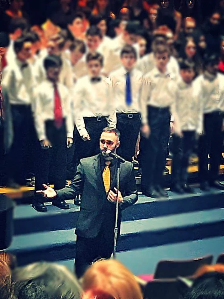 A man in a suit and yellow tie speaking into a microphone in front of a choir of children in white shirts and colorful ties.