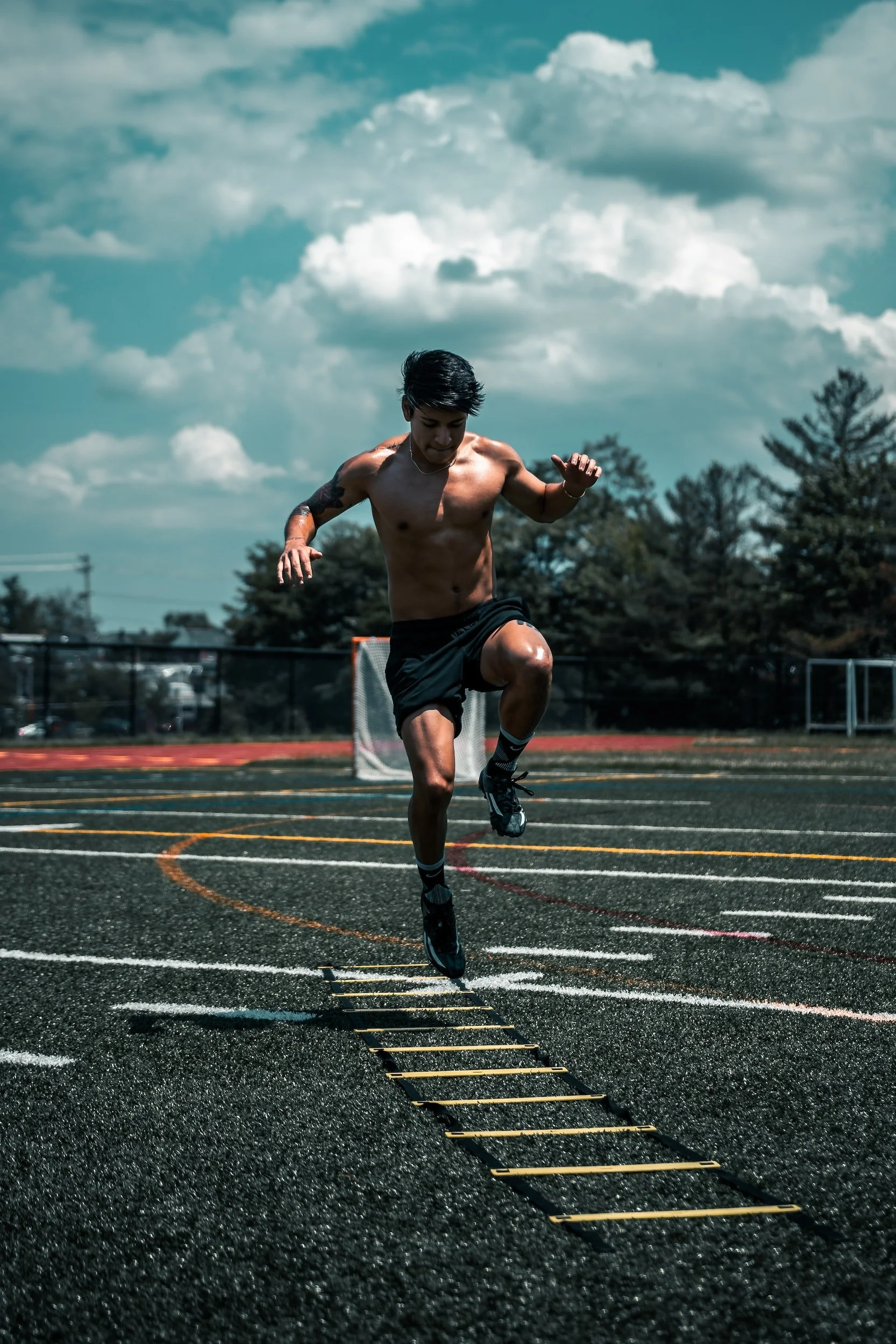 Ein junger Mann trainiert auf einem Sportplatz auf einem plyometrischen Leiter, ist shirtlos, trägt schwarze Shorts, Socken und Laufschuhe, und führt ein Sprungtraining durch.