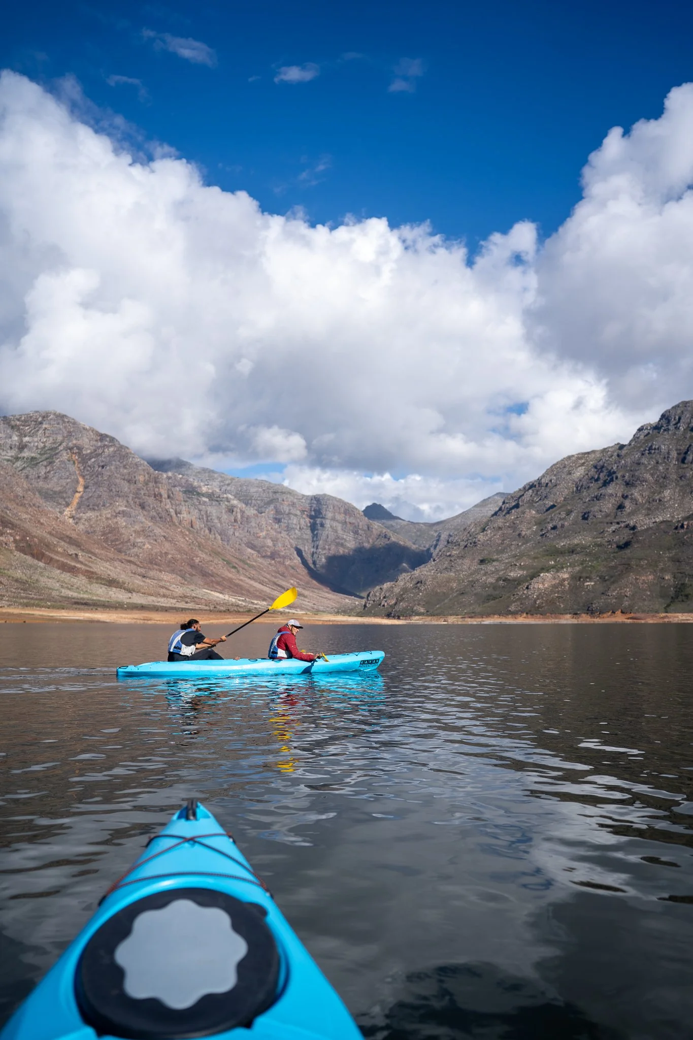 Person kayaking on calm water at sunrise, wearing a hat and life vest, with mountains and the sun in the distance.