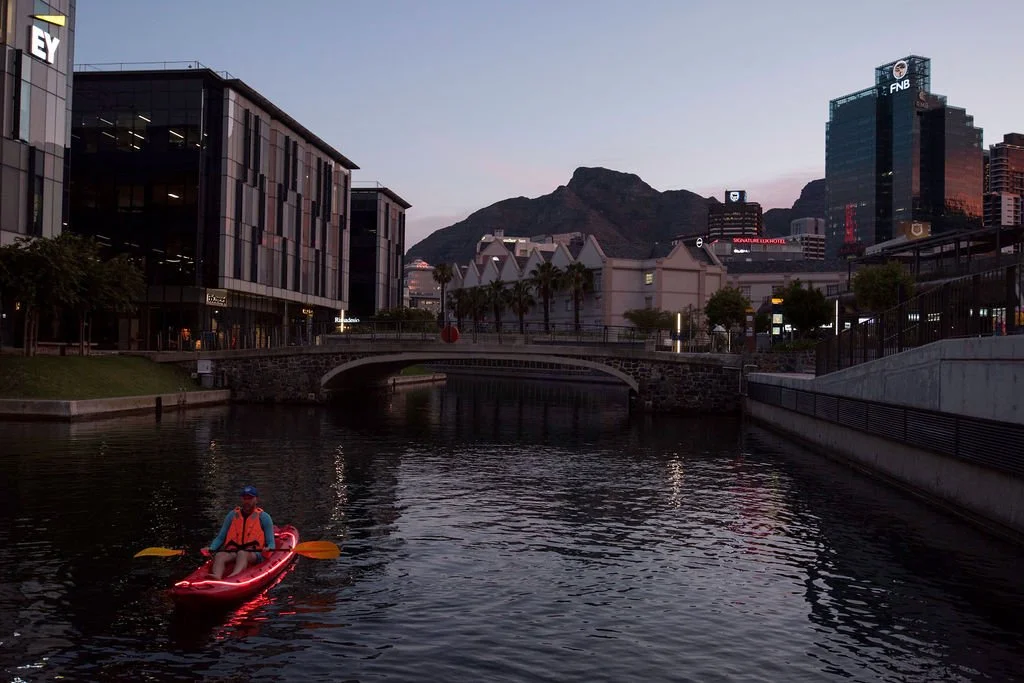 Person kayaking on a canal at dusk with modern buildings and a mountain in the background.