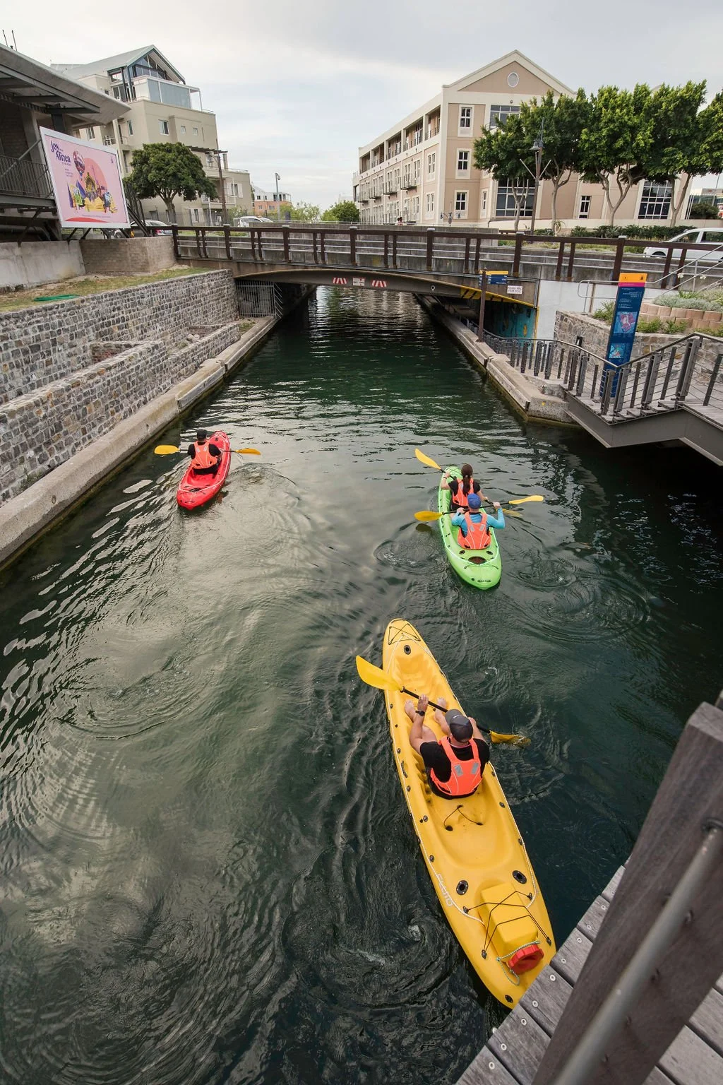 People kayaking through a canal under a bridge, with buildings and greenery on the sides.