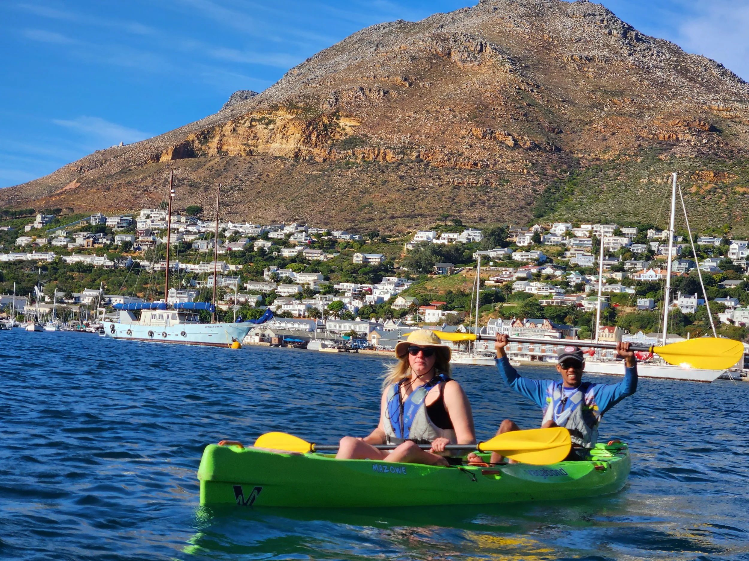 Two people kayaking on calm blue water with a mountainous backdrop, showcasing a scenic coastline and boats docked along the shore.