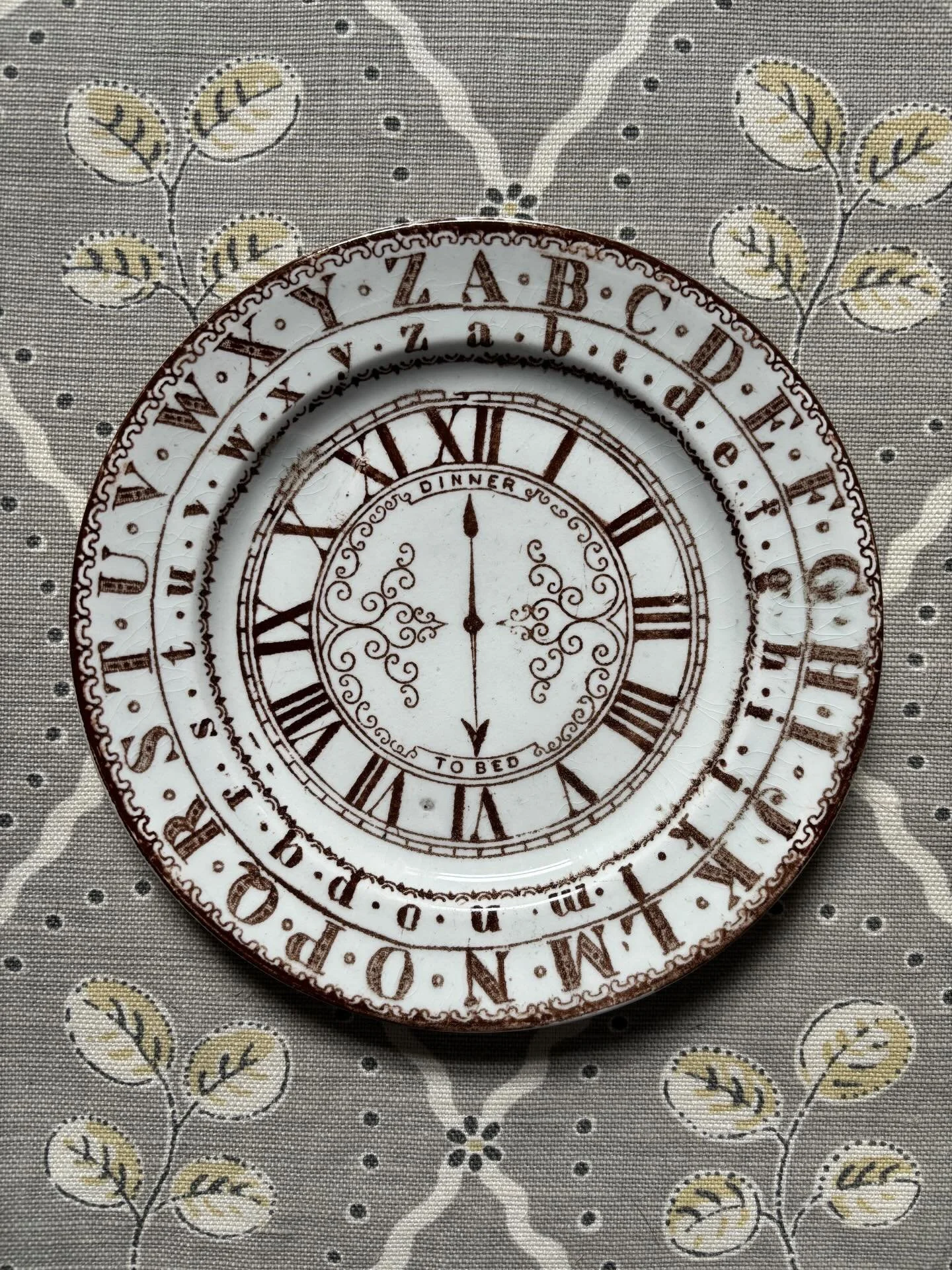 FOR SALE An antique Staffordshire pottery nursery plate, early to mid 19th century, dark brown transfer printed. The hands of the clock point to &lsquo;Dinner&rsquo; and &rsquo;To Bed&rsquo;, the Roman numerals and encircling alphabets signalling its