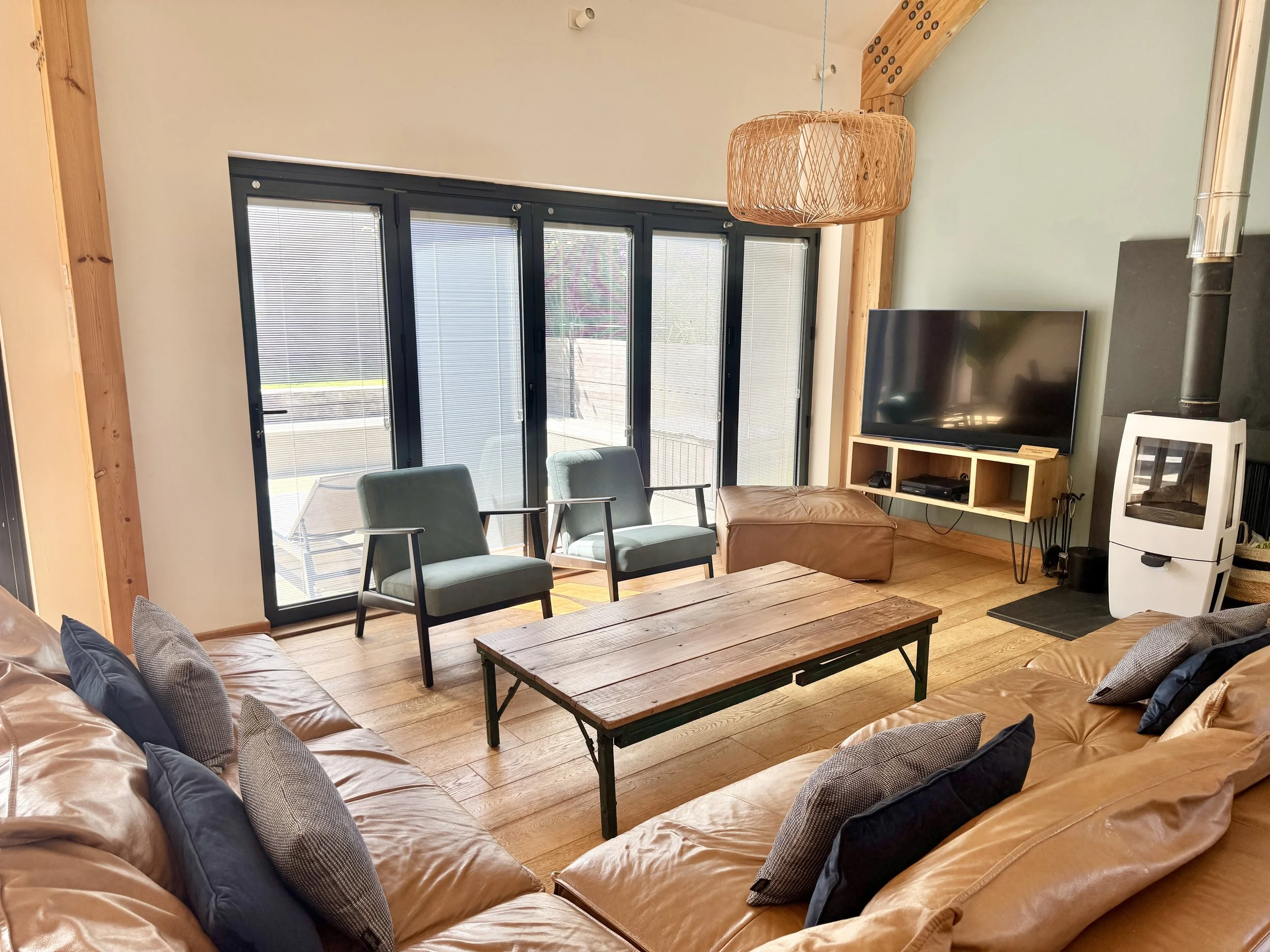 Living room with leather couches, a wooden coffee table, two green chairs, a TV on a wooden stand, a white stove, large glass sliding doors, and a wicker hanging lamp.
