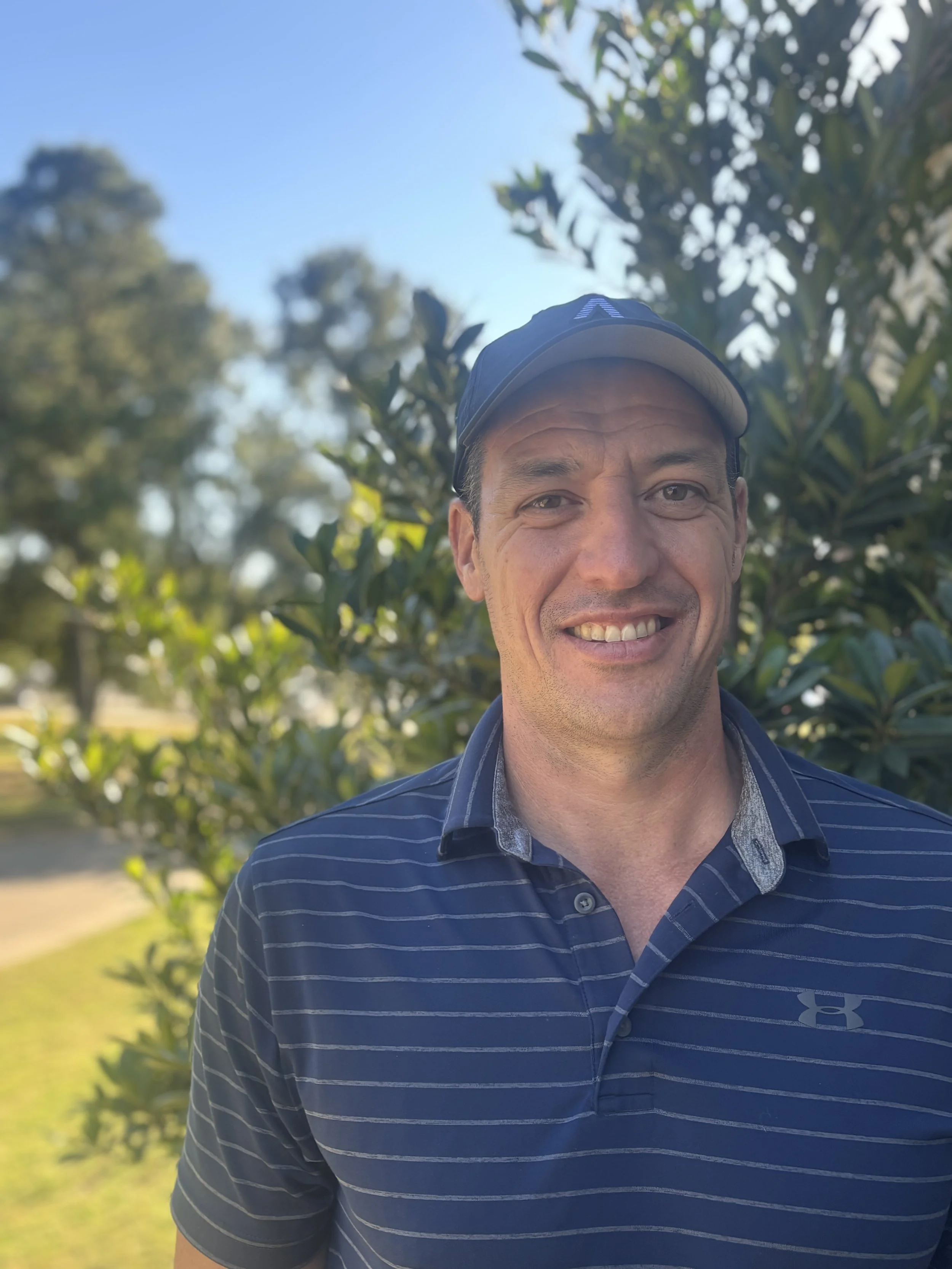 Smiling man in a blue striped Under Armour polo shirt and a cap standing outdoors in front of greenery on a sunny day.