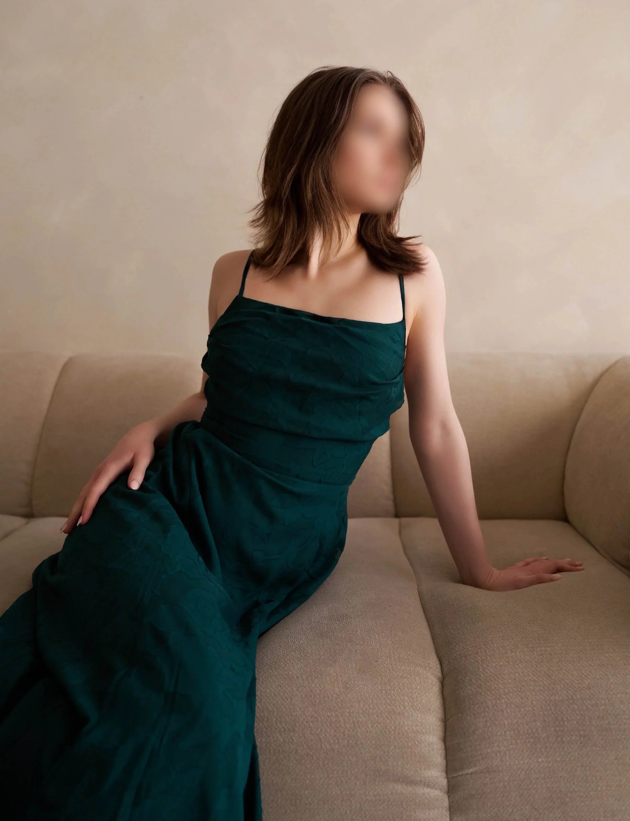A woman with shoulder-length brown hair sits on a beige couch, wearing a dark teal strap dress. She is posed with her right hand on her lap and her left hand on the couch, facing slightly to the right. The background is a plain, light-colored wall.