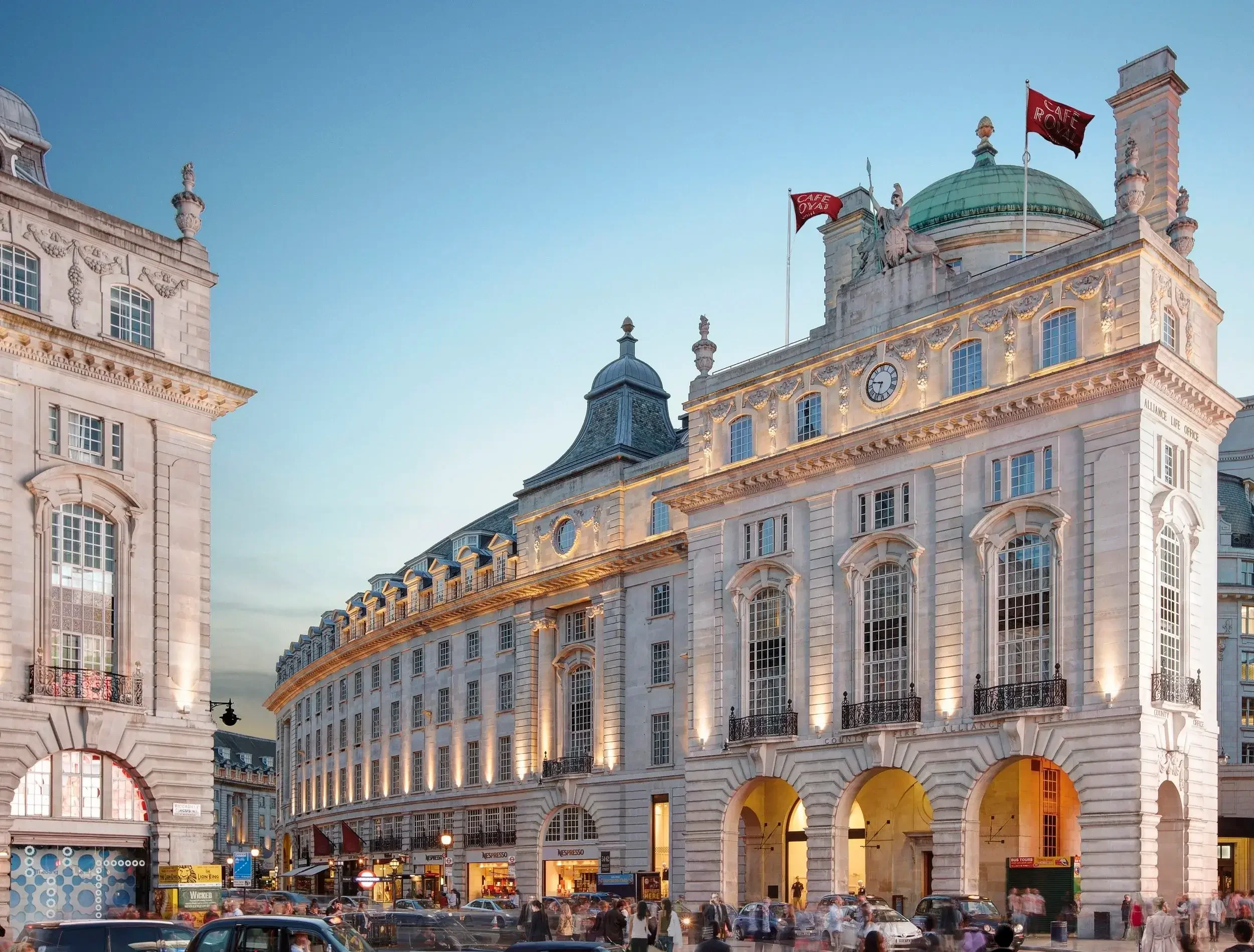 A busy city street scene with elegant historic buildings, cars, and pedestrians during sunset.