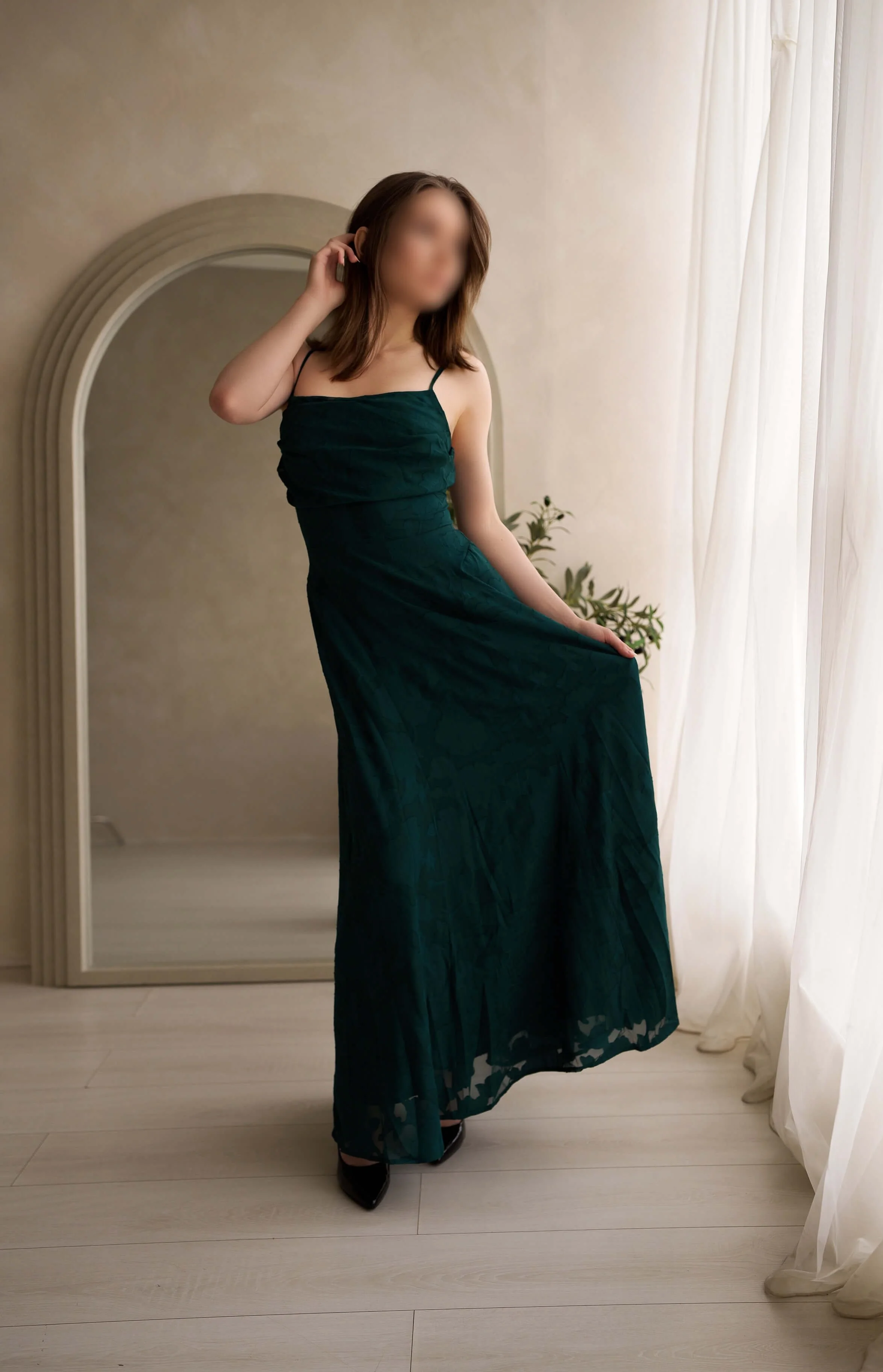 A woman in a dark green dress with spaghetti straps standing in front of a mirror, partially pulling up her dress, with her face blurred. She is indoors near a window with sheer curtains, and there is a small plant in the background.