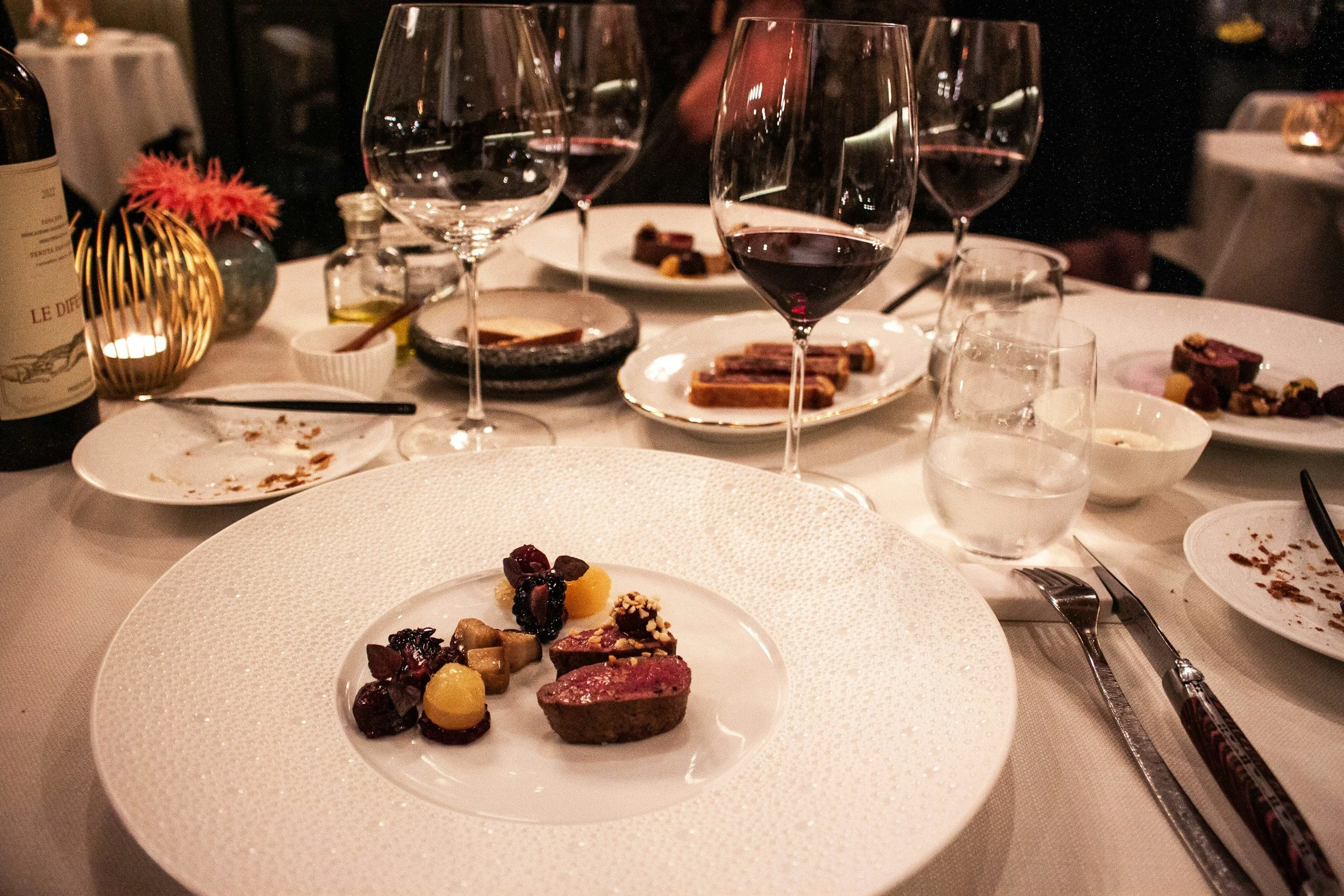 Elegant dinner table set with plates of dessert, glasses of red wine, water, and a small decorative flower arrangement.