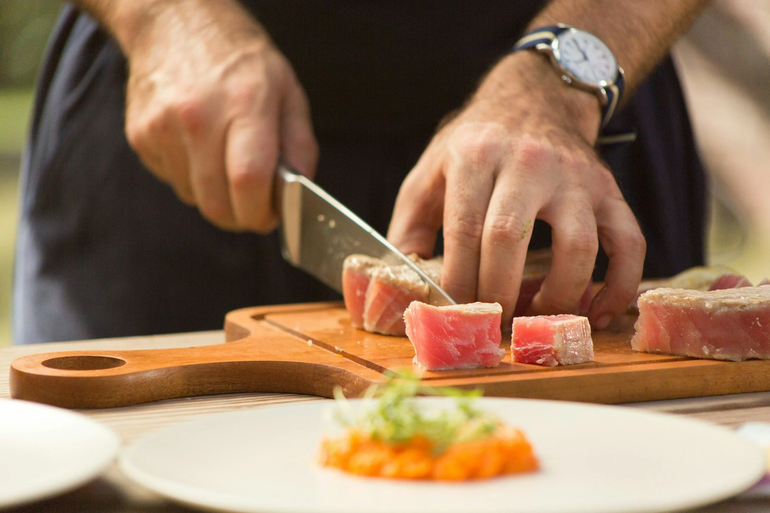 Person slicing raw meat on a wooden cutting board in a kitchen.