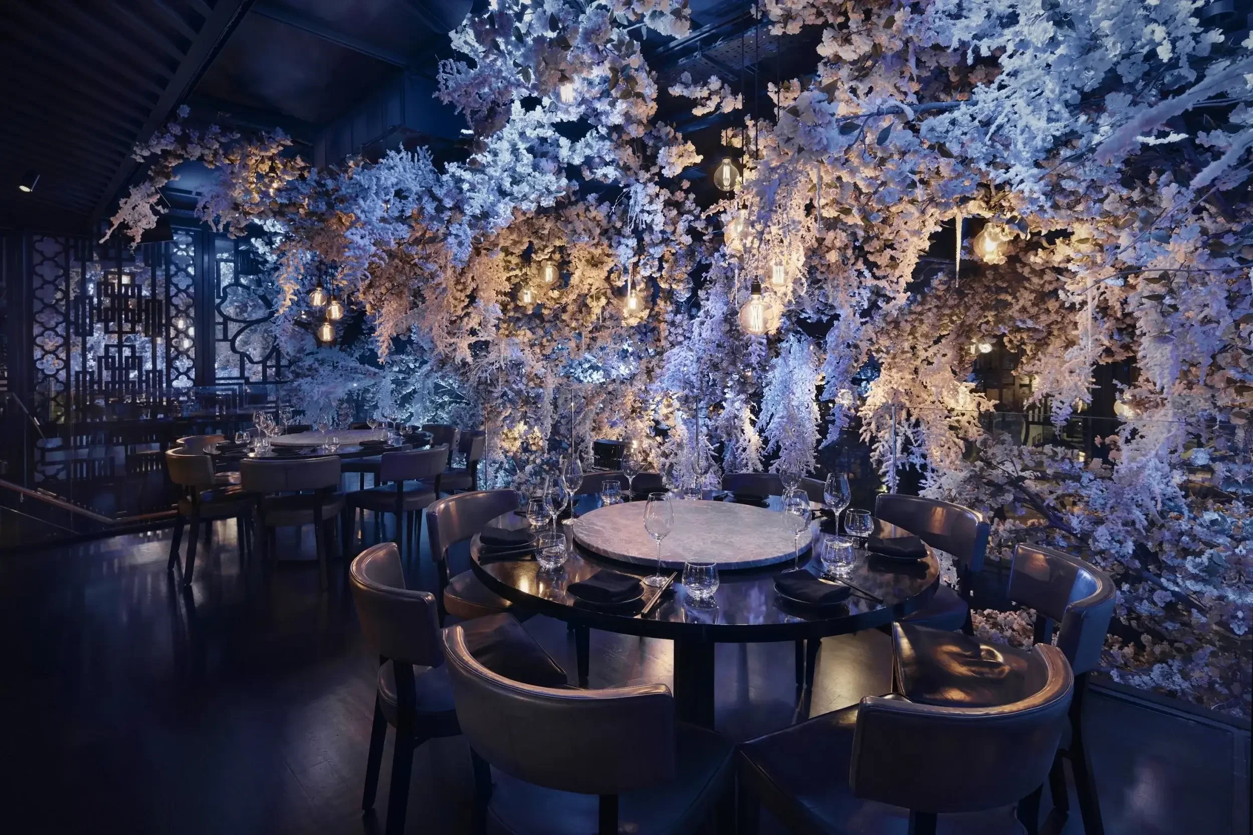 Elegant dining room with round tables, black chairs, and a decorative wall of white illuminated flowers and branches.