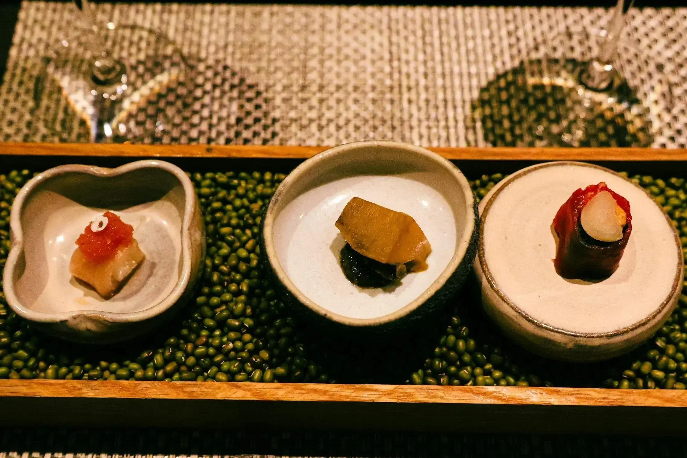 Three small dishes with different types of sushi on a bed of green beans, on a wooden tray, and a placemat with two wine glasses in the background.