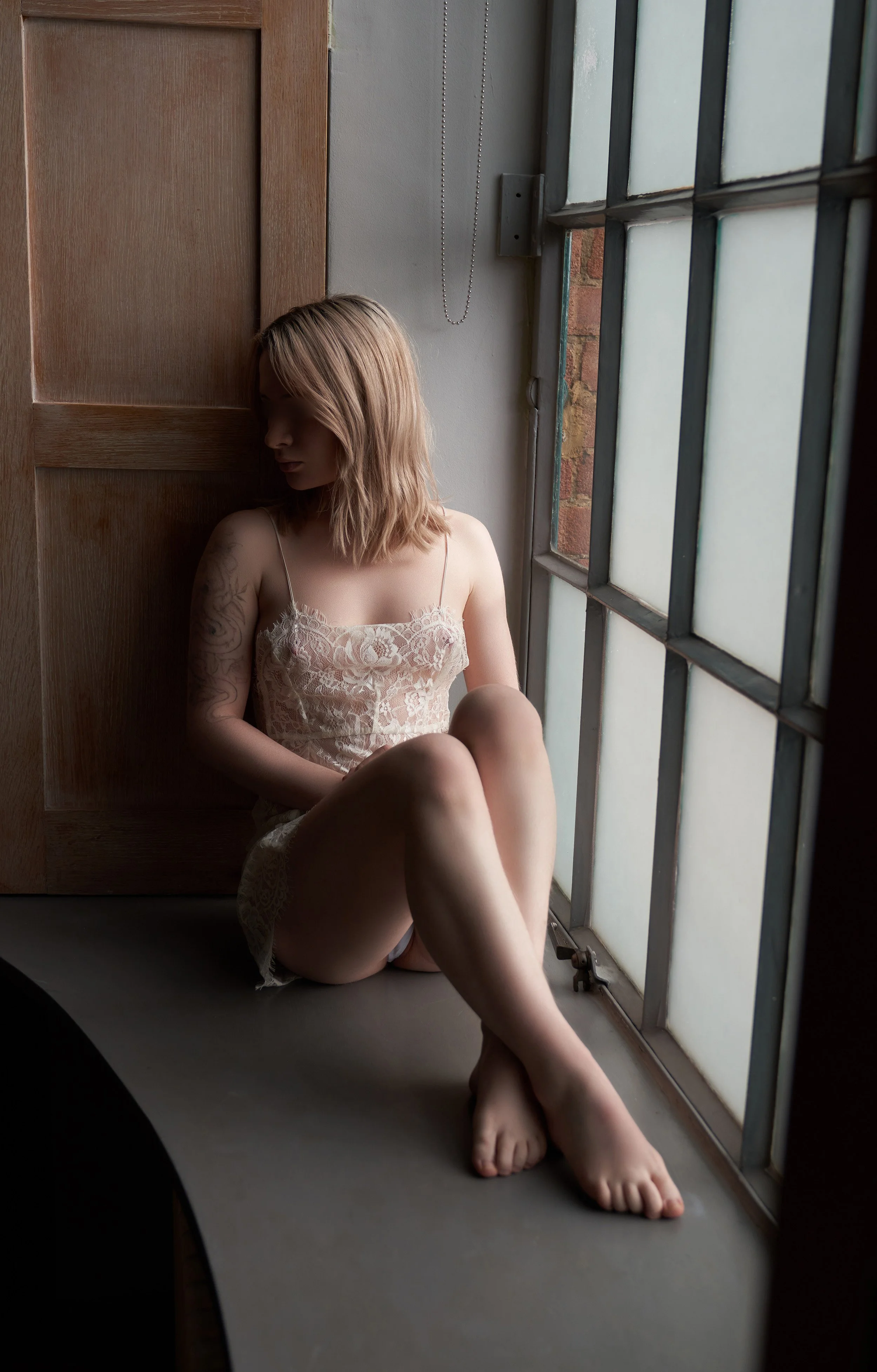 Young woman with shoulder-length hair, tattoo on her upper left arm, sitting on the floor by a window in a lace dress with thin straps, looking down, softly lit by natural light.