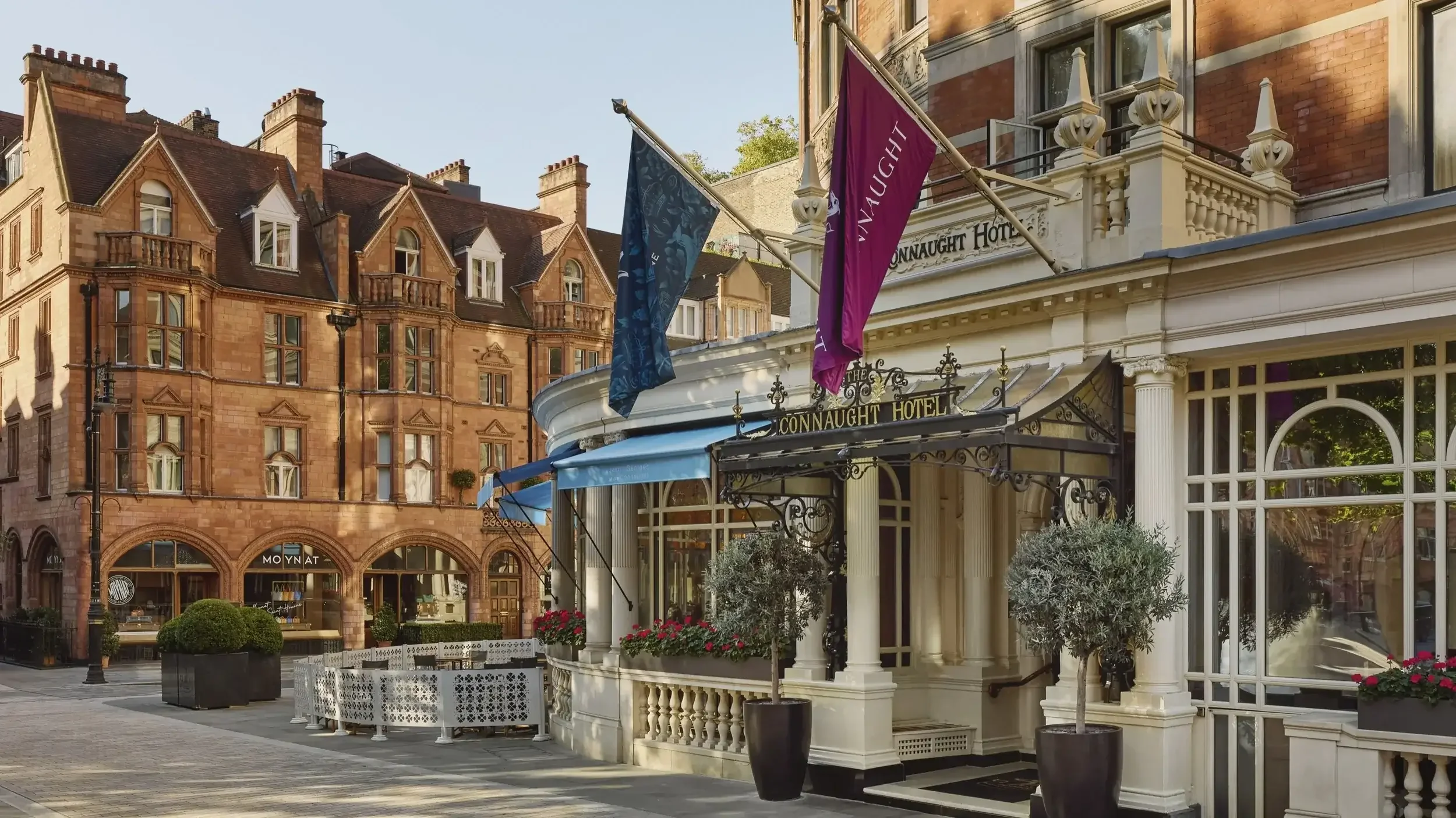 Exterior view of a hotel with white facade and black wrought-iron entrance canopy, potted plants, and flags, in a city street with red brick buildings in the background.
