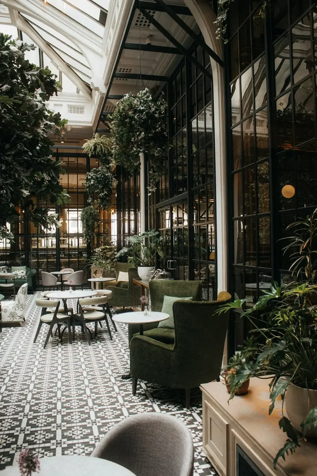 Interior of a stylish cafe with patterned black and white tile floor, green velvet armchairs, white marble tables, and large glass windows with black frames, decorated with hanging plants and lush greenery.