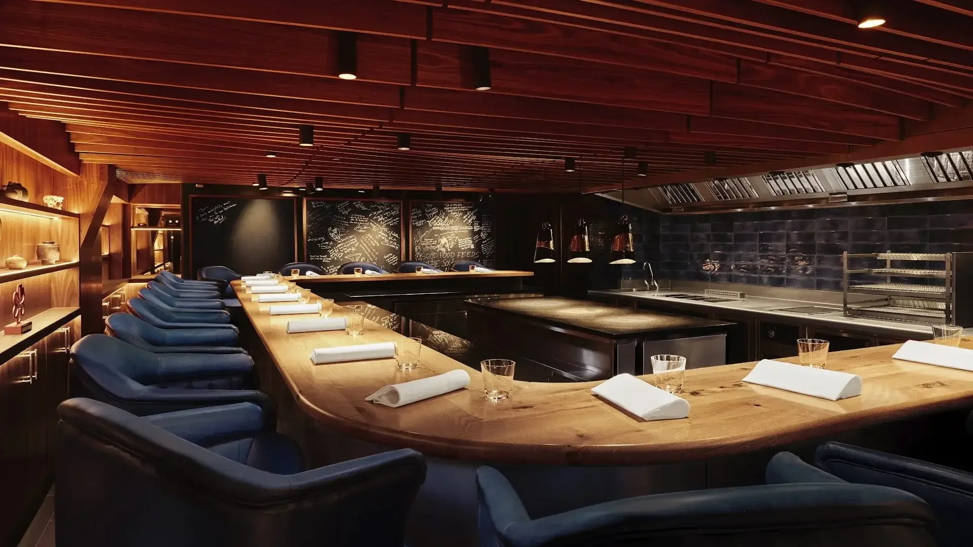 A dimly lit restaurant or bar with a U-shaped wooden bar counter, blue chairs, and shelves with decorative items. There are napkins and glasses on the counter, and a blackboard with writing in the background.